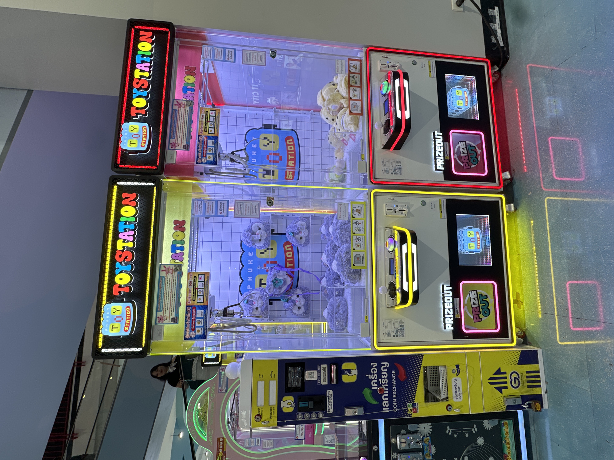 A wide shot captures two brightly lit "Toy Station" claw machines positioned side-by-side in an indoor commercial space, likely a shopping mall, in Wichit, Thailand. The machines are adorned with colorful signage and illuminated displays showcasing plush toys within their glass enclosures. The left machine is predominantly yellow, while the right is pink, both featuring prominent "TOY STATION" logos.
The machine on the left has a "Coin Exchange" section with a basket and instructions. The machine on the right has a "Prize Out" display. The floor beneath the machines reflects the neon lights, creating geometric patterns of yellow and pink. A person, a young woman wearing a dark top and white scarf, is visible in the background, standing on an elevated walkway and looking towards the machines. The overall environment appears clean and well-lit, typical of a retail or entertainment venue.