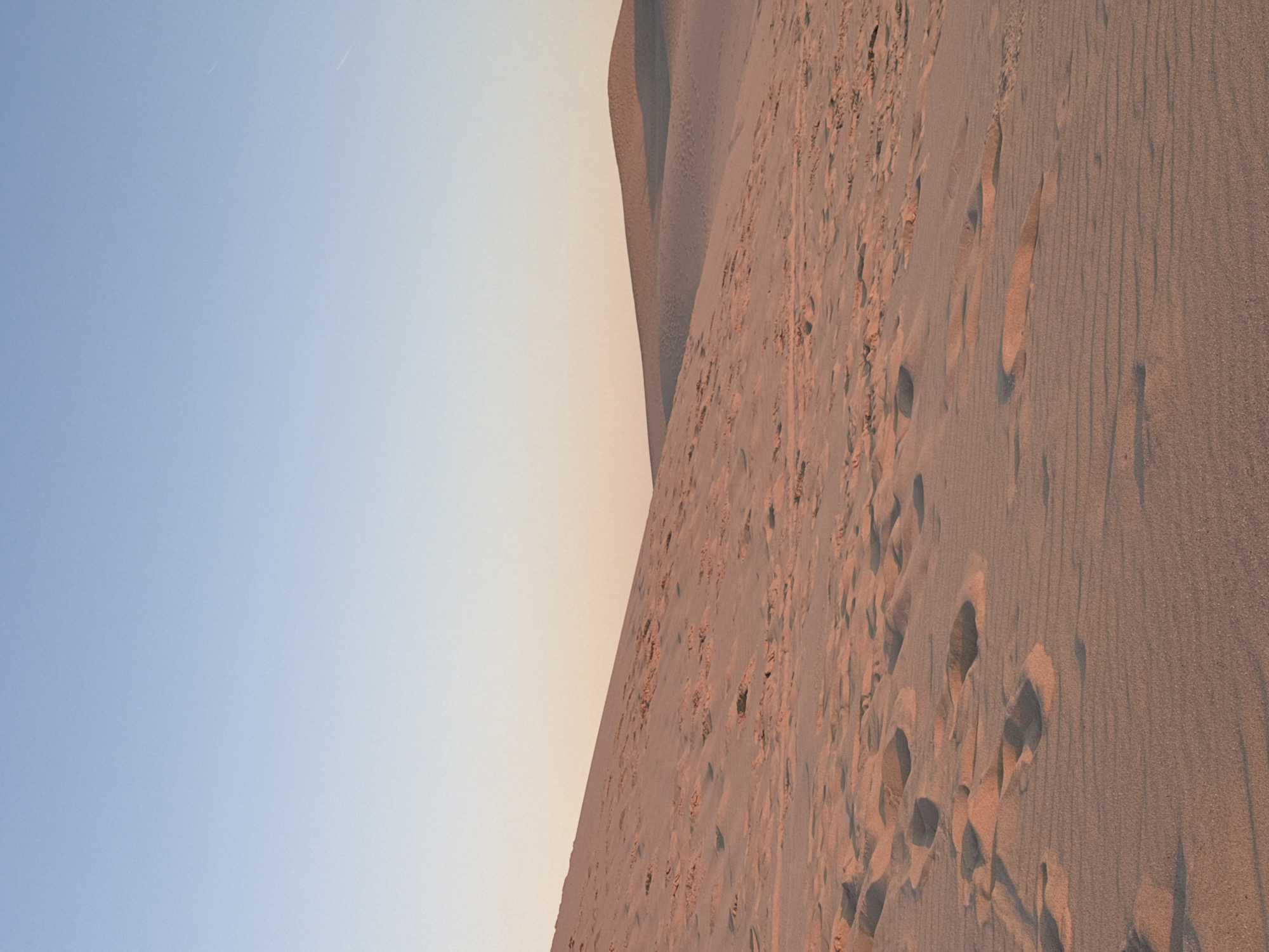 The image depicts a sandy landscape, likely sand dunes, under a clear sky with a gradient from light blue at the top to a pale peach color near the horizon. The foreground and midground are dominated by sand, showing ripples and numerous footprints. In the distance, the landscape curves into rolling sand dunes, casting soft shadows. The lighting suggests it is either sunrise or sunset, with the warm tones illuminating the sand. The location is the Dune du Pilat in La Teste-de-Buch, France, a prominent natural landmark known for its vast sand dunes.