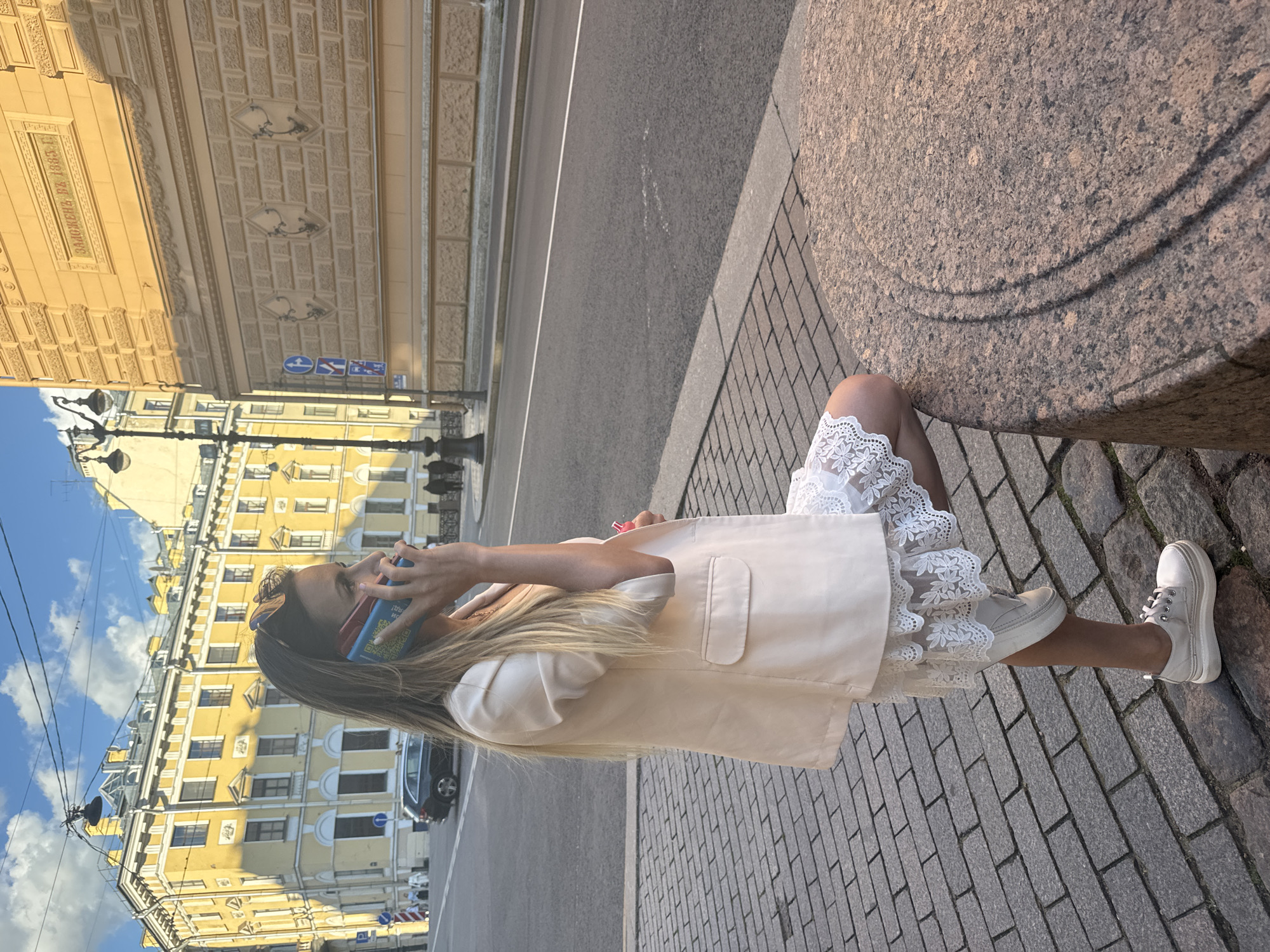 A woman is standing on a cobblestone sidewalk next to a street in Saint Petersburg, Russia, while talking on her cell phone. She is wearing a light beige blazer over a white lace-trimmed dress and white sneakers. Her right hand is holding the phone to her ear, and her left hand is resting on the phone. She is leaning against a round, reddish-brown granite structure. In the background, there are yellow buildings, a black car, a lamppost, and a street with traffic signs. The sky is blue with some clouds.