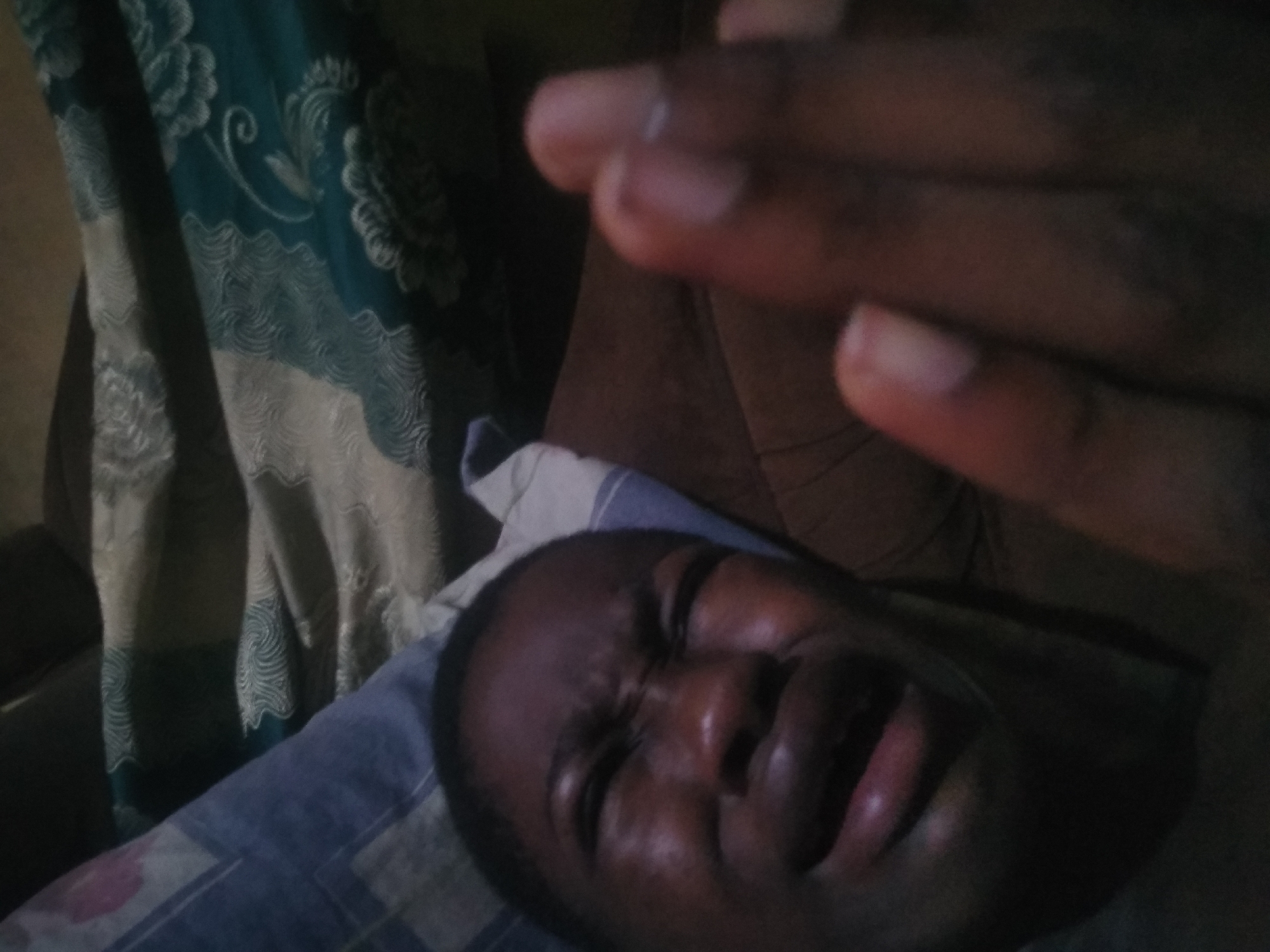 A close-up, low-light indoor shot shows a dark-skinned person lying on a blue and white patterned pillow, possibly in a bed. The person's eyes are closed, and their face appears contorted, possibly in discomfort. A dark-skinned hand is partially visible in the foreground, slightly blurred. In the background, there's a dark floral curtain. No elements are apparent that specifically identify the city of Shangisha, Nigeria.