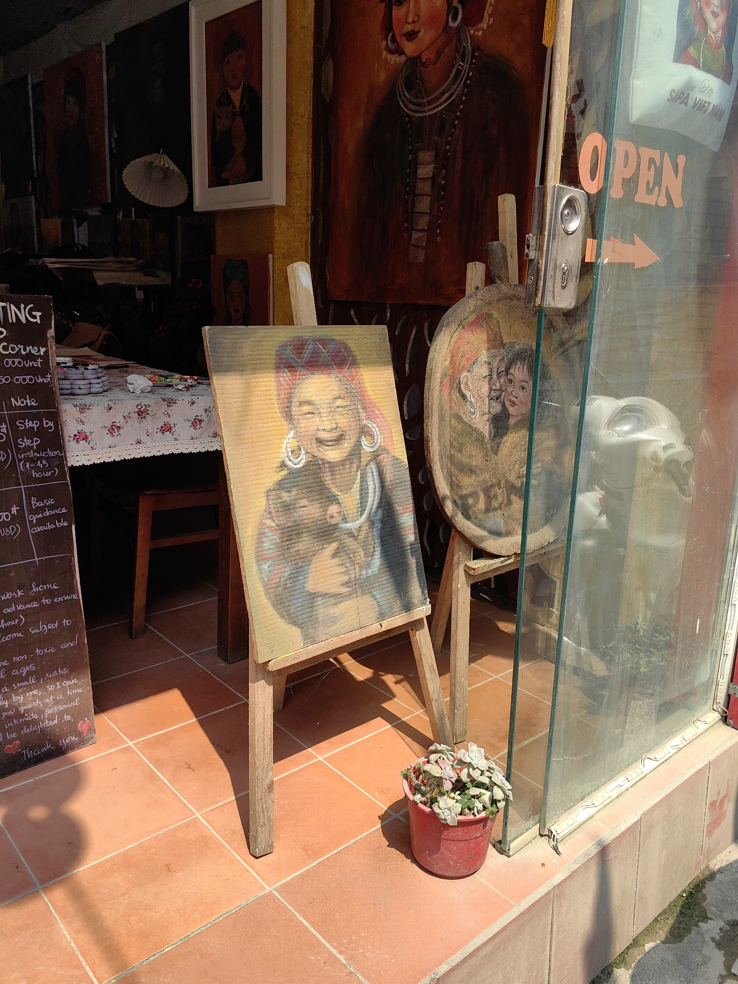 The scene depicts the entrance of an art studio or shop in Sa Pa, Vietnam, identified by text visible through the glass. The foreground features an orange-tiled floor partially illuminated by sunlight. Two wooden easels stand prominently. The easel on the left displays a rectangular portrait of a smiling woman in traditional attire, adorned with large hoop earrings and a distinctive headdress, holding a small, dark animal. The painting exhibits horizontal texture lines. The easel on the right holds a circular painting depicting three faces, two older and one younger, rendered in warm tones. A small red pot containing succulents sits near the base of the right easel.
To the right, a glass door or window displays an orange "OPEN" sign with a directional arrow and a metal locking mechanism. Reflections in the glass show a white scooter or motorbike and a white canvas bag with "SAPA VIET NAM" written on it. A strip of white LED lights is affixed to the lower frame of the glass.
To the left, a black chalkboard sign advertises a "PAINTING CORNER," listing prices in VND and USD, and mentioning "Step by Step instruction" and "Basic guidance." Behind the easels and visible through an opening, an interior space reveals a table covered with a white floral tablecloth, set with small white plates or saucers and painting supplies. A wooden chair is positioned under the table. The walls of the interior are adorned with various framed and unframed paintings, predominantly portraits, including a large portrait of a woman in traditional dress directly behind the right easel. A white, scallop-edged decorative object, possibly a fan or lampshade, hangs in the background.