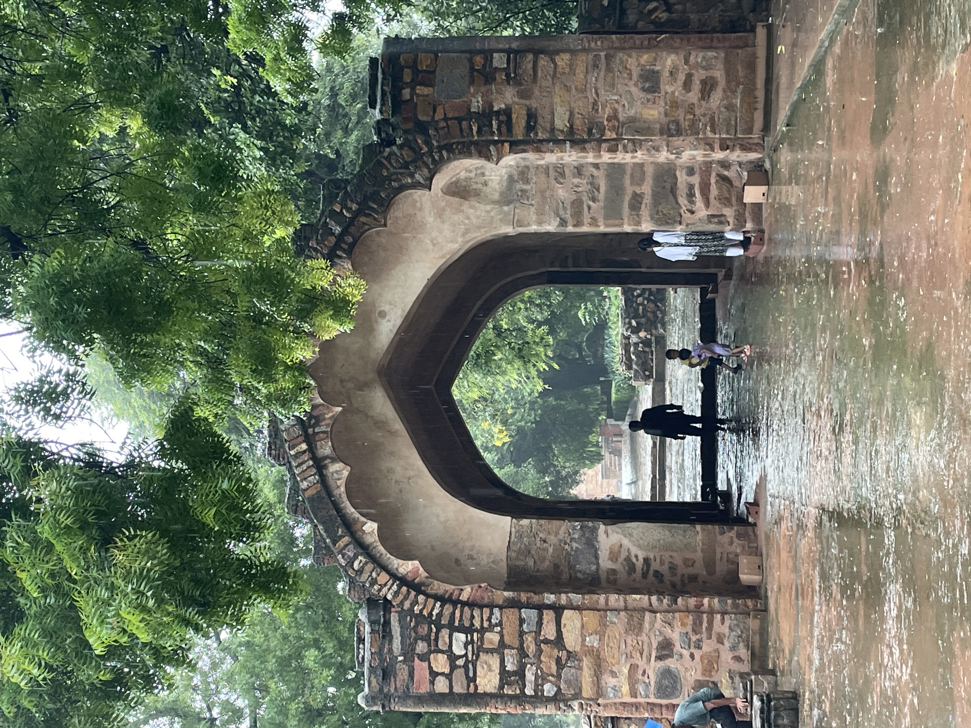 The image depicts an overcast day with rain falling, as evidenced by the water on the ground and visible raindrops. A large, ancient stone archway stands prominently, constructed from rough-hewn stones in various shades of brown and orange, with a lighter, smoother stone arch. The archway is flanked by thick stone pillars. Underneath the arch, a path leads into a courtyard, which also appears wet.
In the foreground, a person wearing a green t-shirt and blue headwear is crouched down on a stone structure, partially visible. To the right of the archway, two children are standing, one in a yellow and purple outfit, and the other in a white outfit with a patterned garment. Further inside the archway, a lone figure, silhouetted against the lighter background, walks away from the camera.
The environment is lush and green, with dense foliage from trees arching over the top of the archway and extending into the background. The ground in front of and under the archway is reflective due to the rain, showing the wet, reddish-brown paving stones. The overall impression is of an old historical site in Delhi, India, possibly a fort or ruin, experiencing a rain shower.