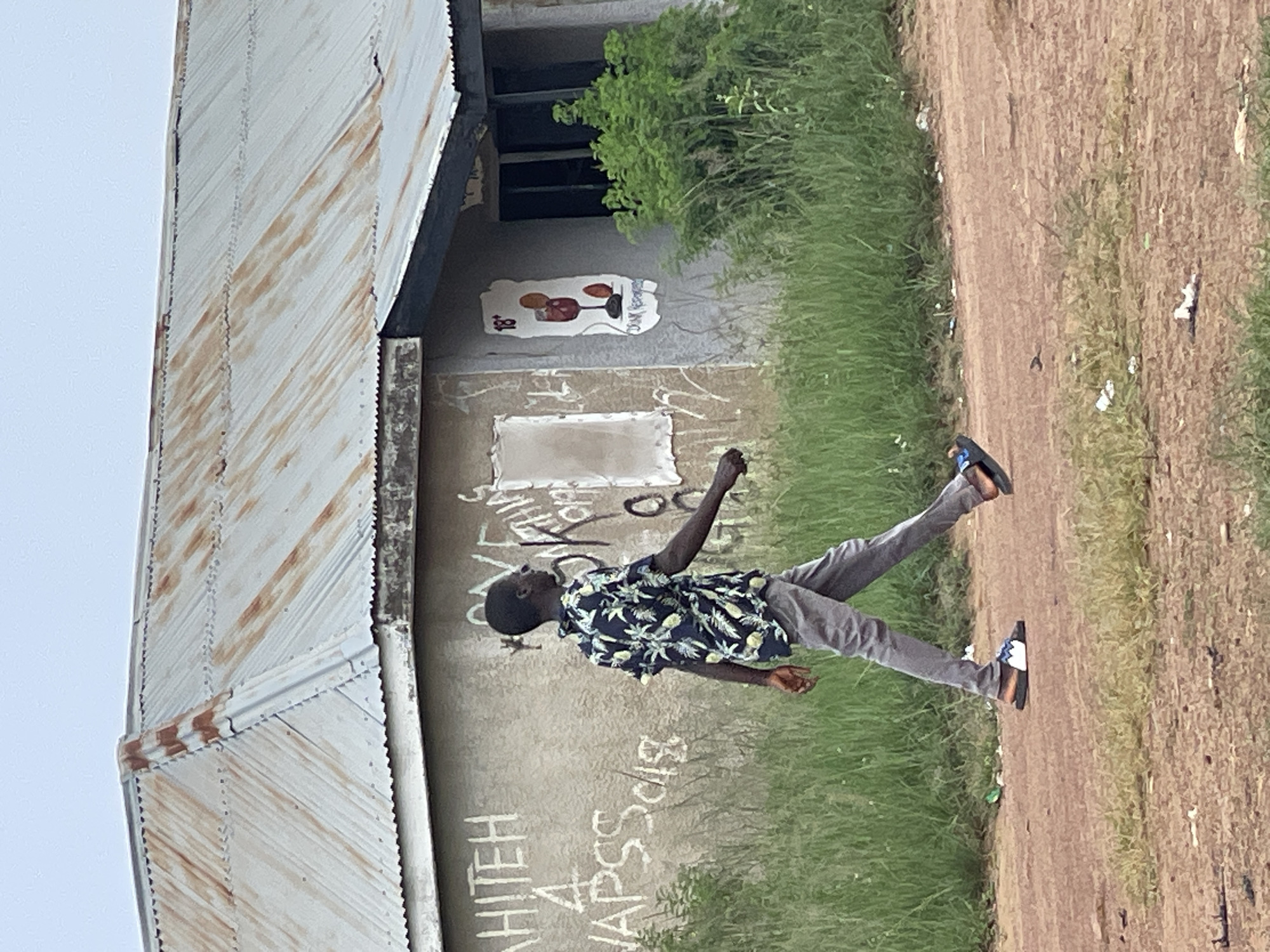 The image shows a young Black male standing in front of a building with a corrugated metal roof. He is wearing a dark floral shirt and grey pants. The building appears to be weathered, with some graffiti on its concrete exterior. A small bush is visible growing near the building, and the ground beside it is covered in grass and dirt. The overall scene suggests an outdoor setting, possibly in a rural or less developed area. The time of day appears to be daytime, and the lighting suggests it might be overcast. The young man's posture and the dynamic composition of the image hint at a moment captured in motion, perhaps mid-stride or gesturing. There is text visible on the wall of the building, appearing to be handwritten graffiti, but it is difficult to decipher clearly. There is also a small, decorative plaque on the wall with a drawing of glasses and what look like two fruits. The location is identified as Durimi, Nigeria.