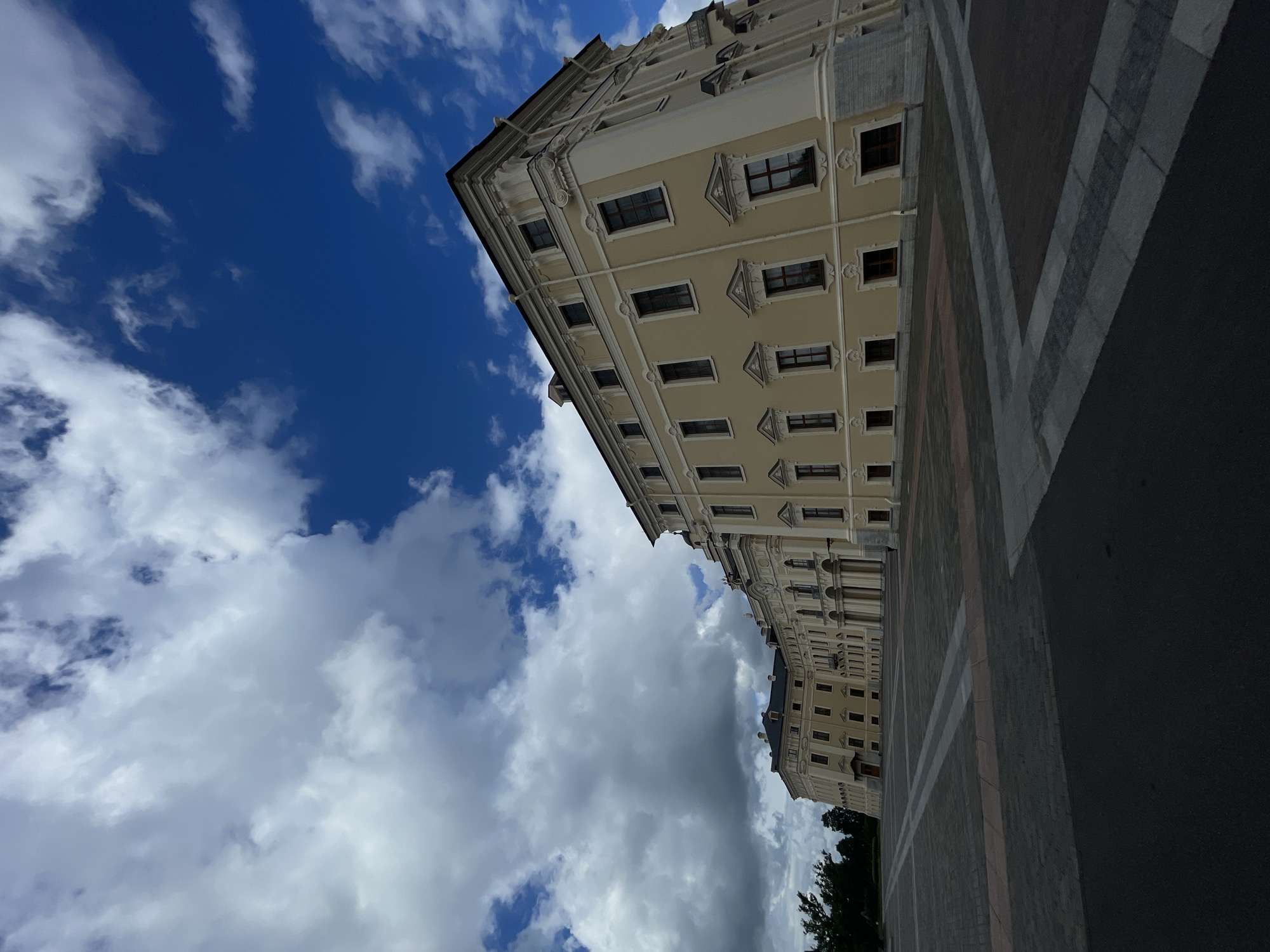 The image depicts a wide-angle view of the Konstantinovsky Palace in Strelna, Russia, under a cloudy blue sky. The palace, a grand yellow building with classical architectural elements, dominates the right and center of the frame, stretching into the distance. Its facade features numerous windows, decorative moldings, and a prominent colonnade. The foreground is a vast, paved courtyard, laid out with patterned stone tiles and dark asphalt. To the left, a few trees are visible, bordering the palace grounds. The sky is a vibrant blue, interspersed with large, dramatic white and grey clouds. There are no people visible in the scene. The perspective is low, emphasizing the scale of the building and the expanse of the sky.