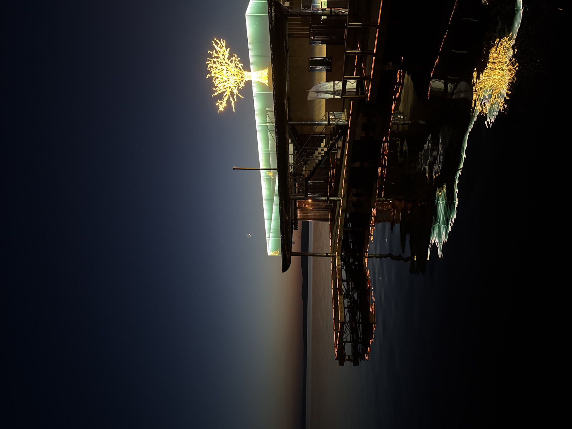 The image depicts a building on a pier reflected in dark water, possibly at night or twilight. The building is illuminated with yellow lights, including a tree-shaped light display on the roof. A crescent moon is visible in the dark sky above the building. The horizon shows a fading sunset or twilight effect. Based on the location hint of Votkinsk, Russia, this may be a pier building on the Votkinsk Reservoir.