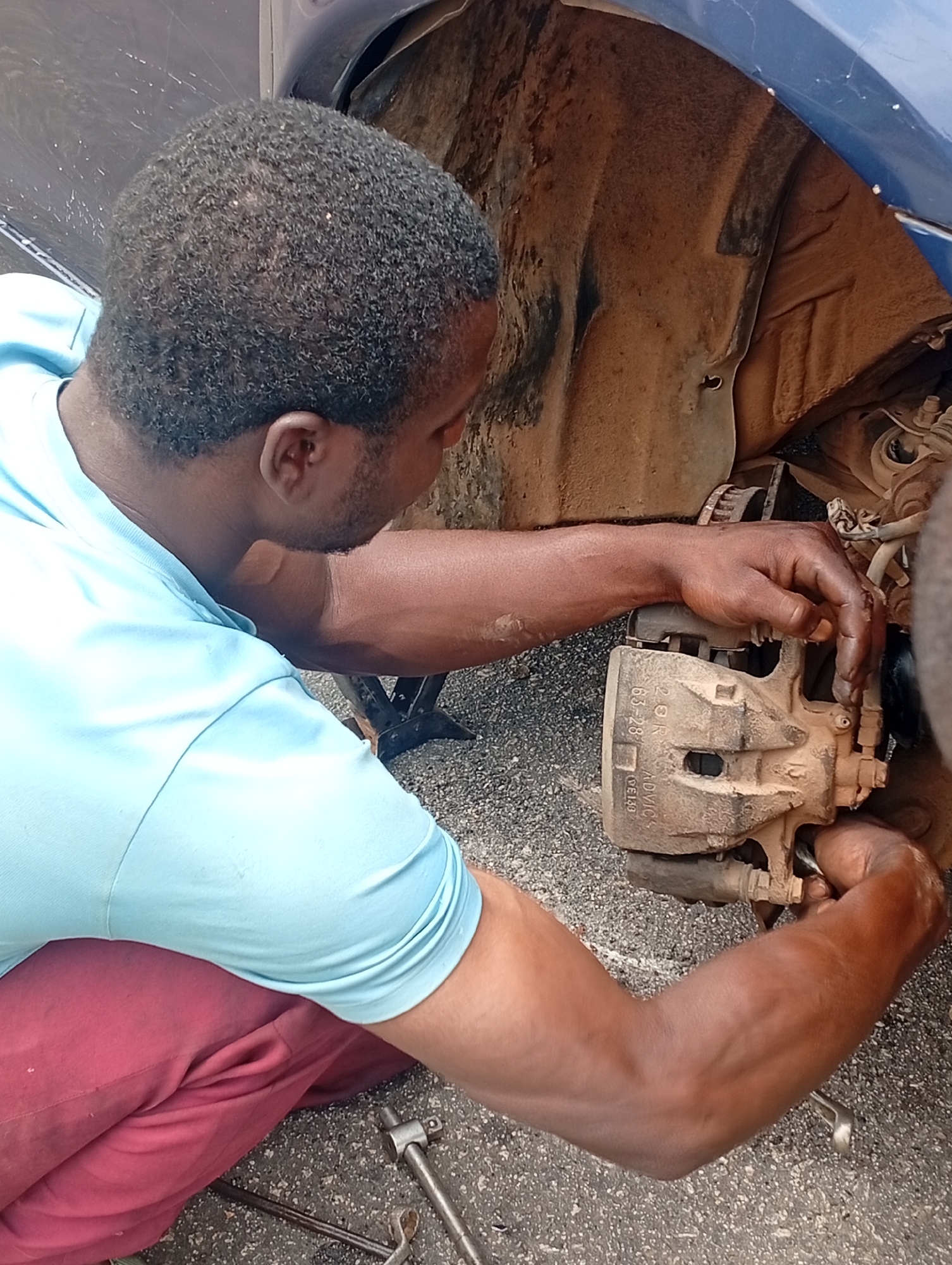 A man is working on the brakes of a blue car. The location is Jos, Nigeria. The car is jacked up, and its wheel has been removed. The man is wearing a light blue shirt and red pants. He is focused on the brake caliper, and tools are lying on the ground nearby. The car's wheel well is dirty.