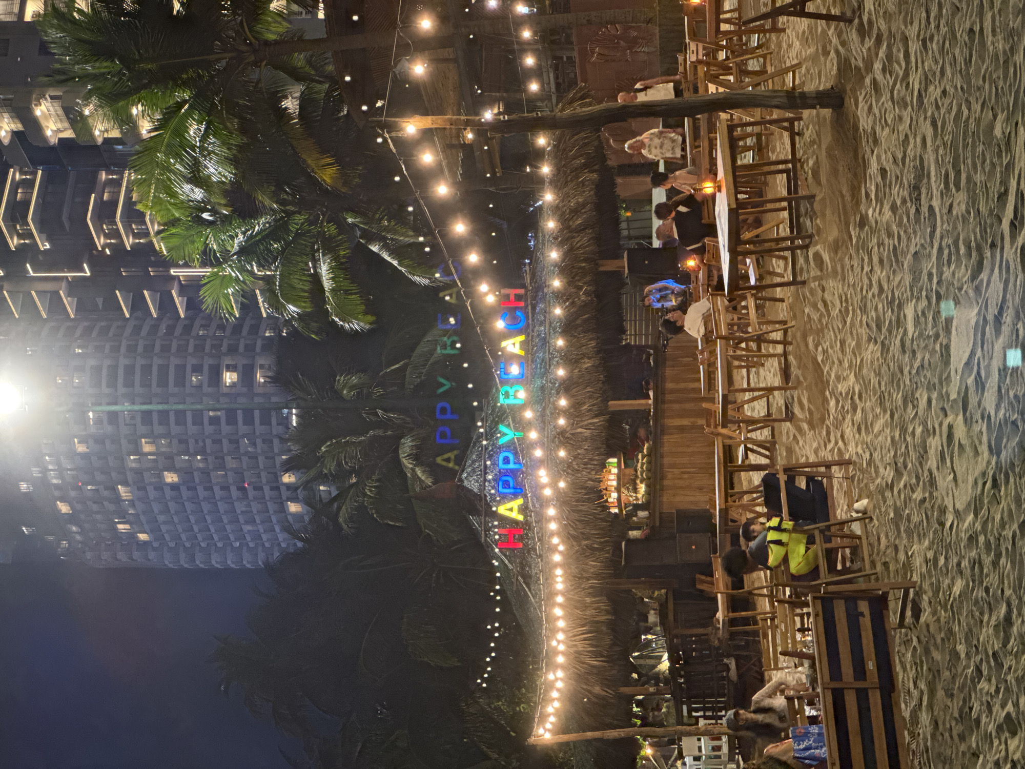 The scene is an outdoor restaurant at night, situated on a beach in an unknown city. The restaurant is adorned with string lights. The words "HAPPY BEACH" are displayed in colorful, illuminated letters. Several people are seated at tables, appearing to dine or converse. The ground is sand. In the background, there is a tall building. Palm trees are also visible.