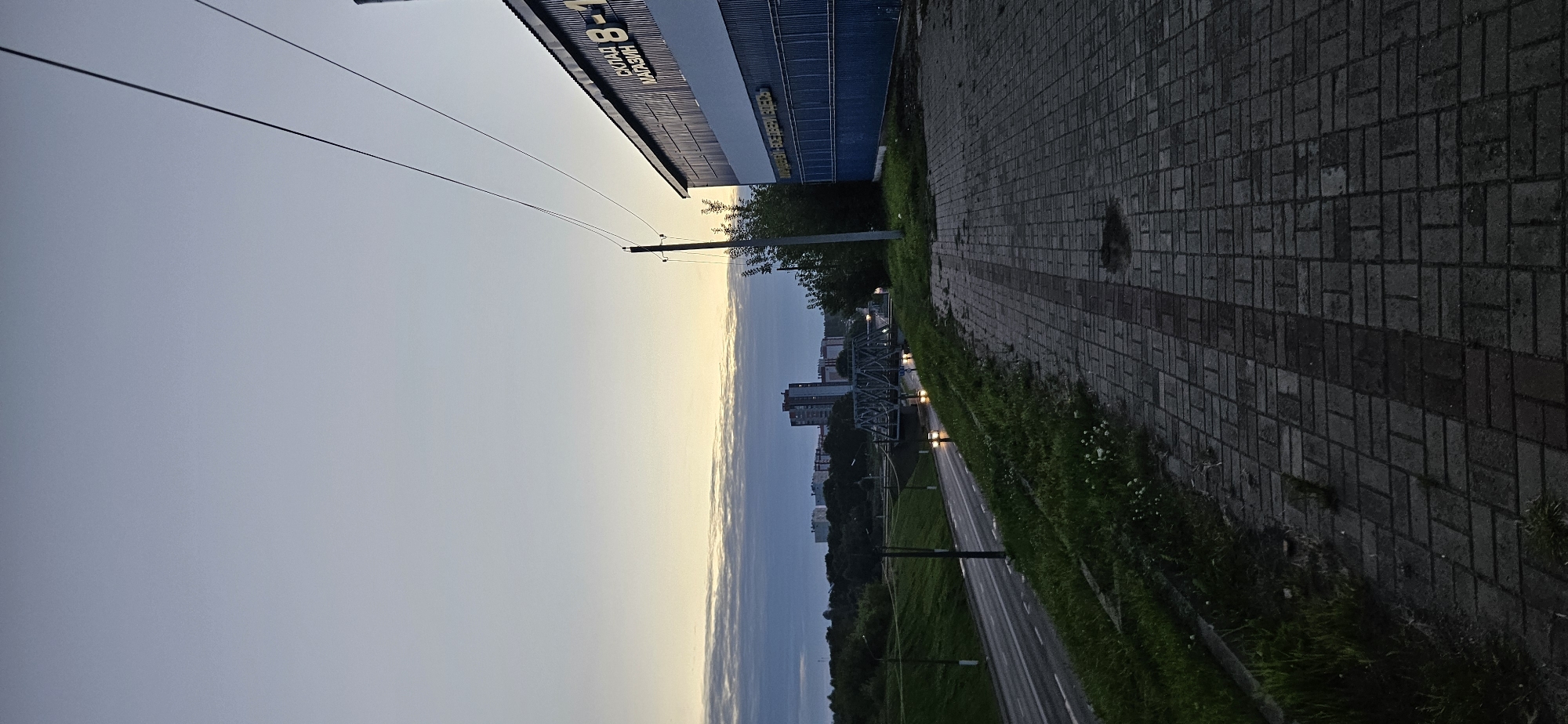 The image depicts a paved walkway in front of a blue and white industrial building with signage that reads "СКЛАД МАГАЗИН 8-". To the left of the walkway, there is a grassy embankment sloping down to a road with cars on it. In the background, a metal bridge spans across what appears to be a body of water, with apartment buildings visible beyond the bridge. The sky is overcast with a hint of sunset colors on the horizon. The overall scene suggests a commercial or industrial area on the outskirts of a city, likely in Vitsebsk, Belarus, given the Cyrillic signage. There are no people visible in the image.