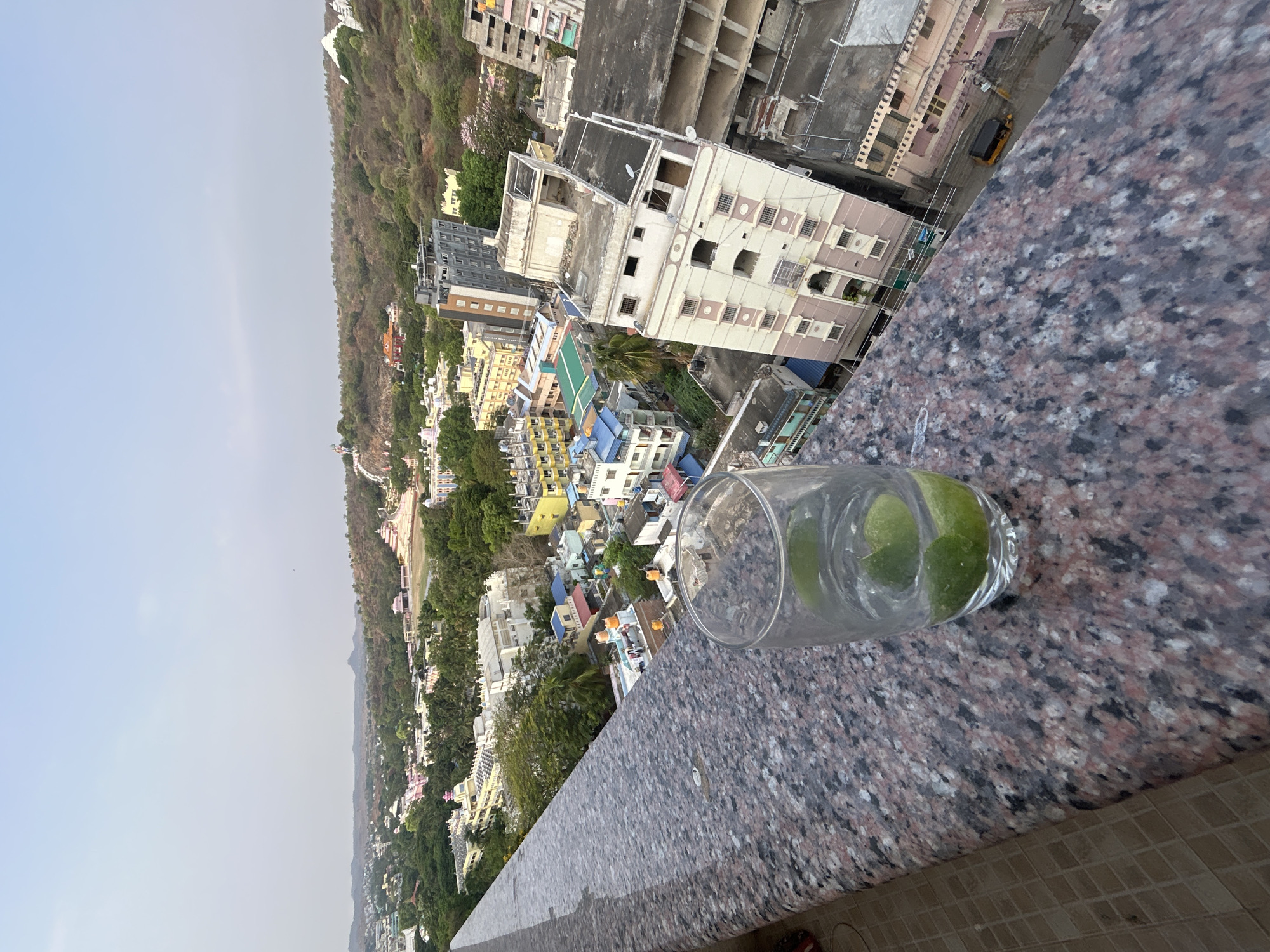 Here is a description of the image:
The photograph is taken from a high-angle perspective, likely from a balcony or rooftop in Puttaparthi, India. In the foreground, a glass of water with slices of lime sits on a stone ledge. The ledge has a mottled dark gray and pink granite-like finish. Beyond the ledge, there is a sprawling cityscape consisting of a mix of low-rise buildings, primarily in yellow, white, and blue. A large hill, covered in greenery, rises in the background to the left, with some buildings nestled into the hillside. The sky is overcast and hazy. In the background, various buildings are visible, ranging from residential to commercial structures. The scene appears to be shot in daylight, although the light is diffused due to the overcast sky. An auto rickshaw sits in front of the buildings in the background.