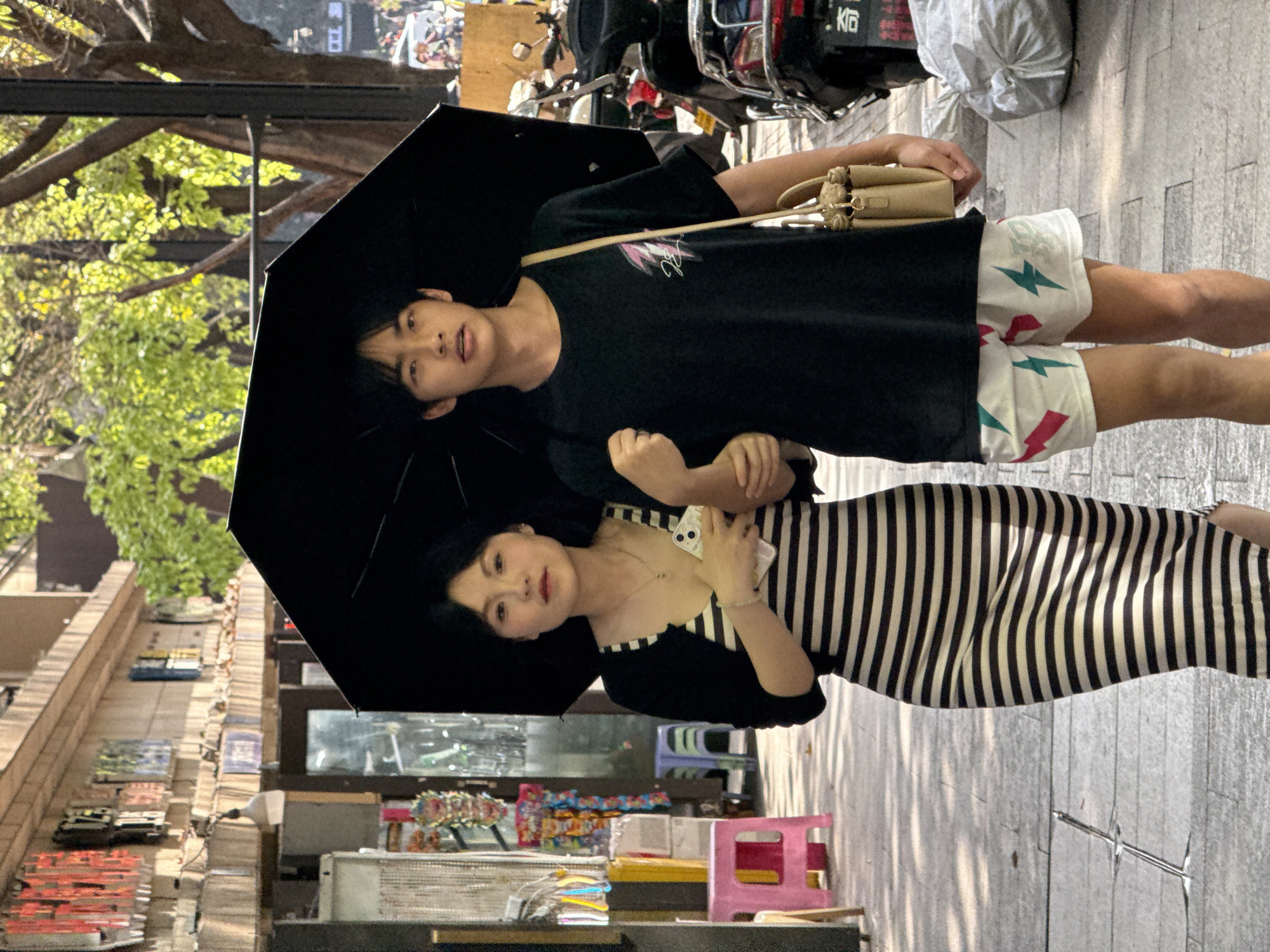 Two individuals, a man and a woman, are captured standing on a paved sidewalk, holding a large black umbrella for shade. The scene appears to be a sunny day in an urban or semi-urban setting, likely in Yuzhong District, China, given the bright lighting and distinct shadows.
The man, on the left, wears a black short-sleeved t-shirt with a faint light-colored graphic and white shorts patterned with teal and red lightning bolts. He carries a beige crossbody bag and holds the black umbrella open above their heads. His expression is neutral as he looks towards the viewer.
The woman, on the right, wears a black and white vertical striped dress, possibly with a black top or cardigan over it. She holds a white smartphone with the Apple logo visible on its back against her chest and has her arm linked with the man's. She also has a neutral expression, looking towards the viewer.
The background shows elements of a city street, including lush green trees on the left, suggesting a park or a treelined area. To the right, behind the man, are parked motorcycles or scooters, with "K6" visible on one. Further in the background are various storefronts displaying merchandise, some colorful, and a pink plastic stool on the sidewalk. The ground is paved with light-colored rectangular blocks, and long shadows are cast by unseen objects, reinforcing the bright, sunny conditions.