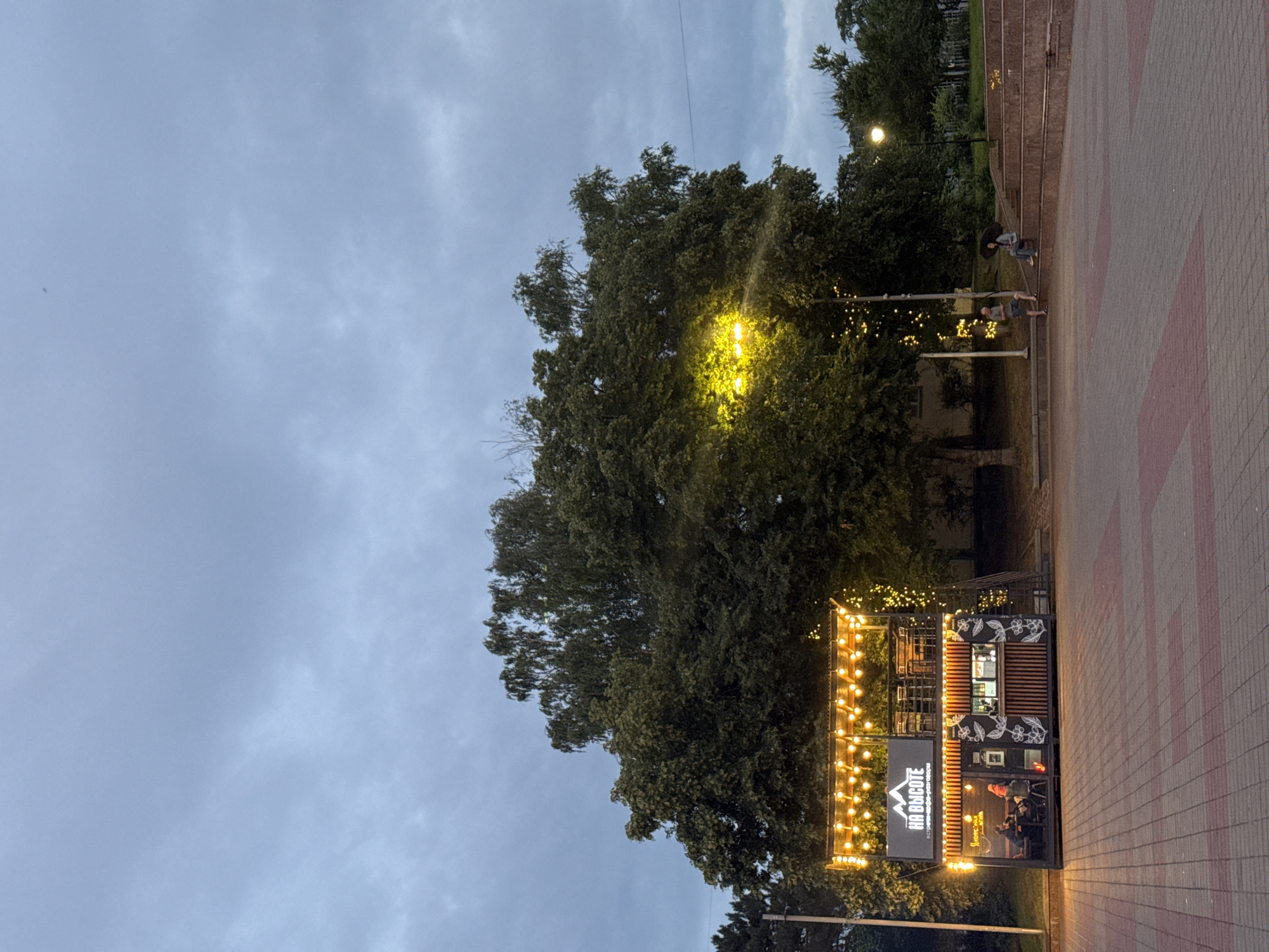 The image depicts an outdoor scene in Mikhaylovsk, Russia, at dusk. In the foreground, a paved area with red and gray interlocking bricks leads to a two-story structure that appears to be a cafe or food stand. This structure is illuminated by strings of warm-toned lights along its upper edge and features a sign that reads "НА ВЫСОТЕ" (Na Vysote), which translates to "At Height." The lower level of the structure has large windows revealing interior seating and a counter area, while the upper level has an open-air seating area with tables and chairs.
Behind the structure, a large, mature tree with dense foliage dominates the mid-ground. To the right of the tree, a set of tiered stone steps forms a seating area where two individuals are visible. One person is sitting with an umbrella, and another is standing nearby. A street lamp casts a warm yellow glow on the tree and the surrounding area. The sky is overcast with a pale gray hue. The overall atmosphere suggests a public space in a city, possibly a park or a town square, during the early evening hours.
