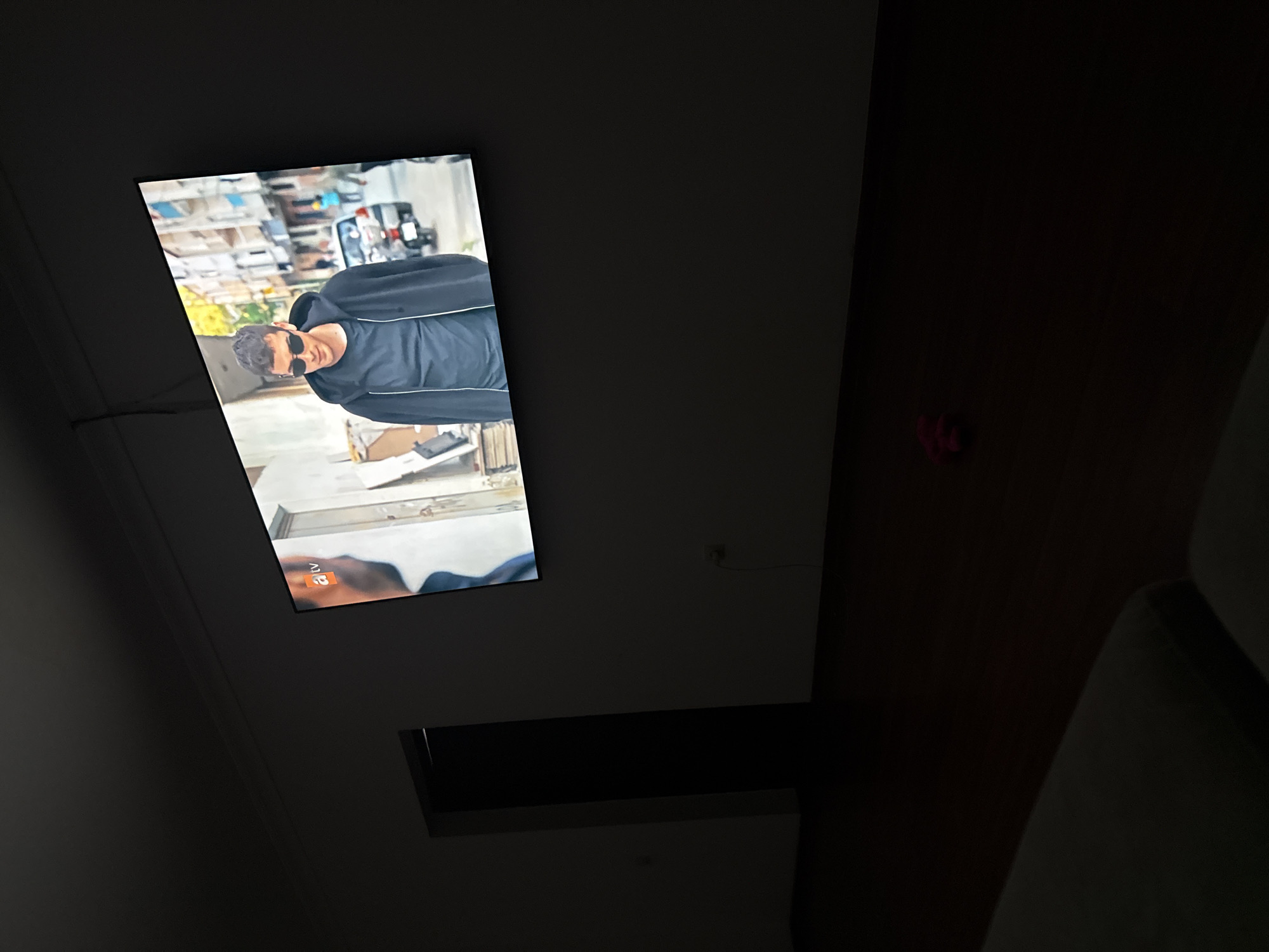 In a dimly lit room, a large flat-screen television displays a scene from a TV show. The show features a young man wearing sunglasses and a black hooded jacket over a dark t-shirt, standing on a city street with buildings and a car in the background. The room itself is sparsely furnished, with a dark wooden floor and a partially visible doorway leading to a darker space. A red object, possibly a toy, is on the floor in front of the television. The overall impression is that of a private residence, likely in the city of Serebryakovka, Russia, where someone is watching television in the evening.
