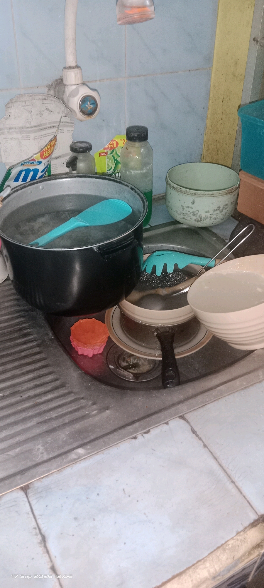 The image depicts a cluttered kitchen sink area, likely in a residential setting in Klaten, Indonesia. A large black pot filled with water and a turquoise rice paddle sits in the sink. To the right of the pot, there are two bowls, one containing a white substance, possibly soap suds or food. A scrub brush with a turquoise handle rests on a plate beneath a second pot. On the counter behind the sink are several bottles, including one of dish soap and a smaller container with a black lid. A damaged piece of paper, possibly a label or instruction manual, is also visible near the faucet. The sink and surrounding surfaces appear to be made of stainless steel and tiles, showing signs of use and potential grime. The overall impression is one of a working kitchen, possibly in the process of or after washing dishes.
