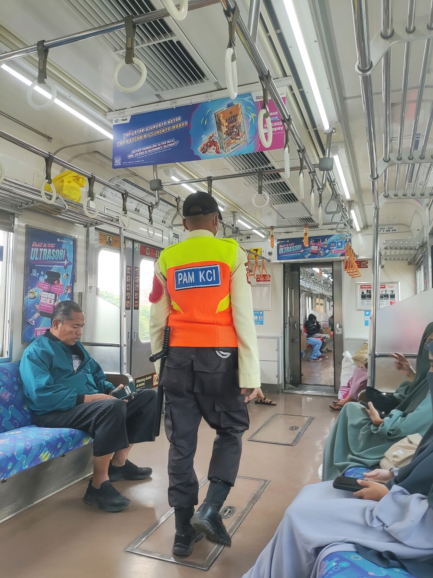 The image shows the interior of a commuter train. In the foreground, a person wearing a high-visibility vest over a long-sleeved shirt and dark pants is walking away from the camera. The vest is orange and yellow, with "PAM KCI" printed on the back in blue letters. They are also wearing a dark cap. To the left, a man is seated on a blue and white patterned seat, looking down at his phone. He is wearing a teal jacket and dark pants. The train car has silver grab bars and handrails overhead, with fluorescent lighting. Advertising posters are visible on the walls. In the background, through an open doorway, other passengers can be seen seated. The overall environment appears to be a standard commuter train carriage. The provided context states the location is Tambun, Indonesia.