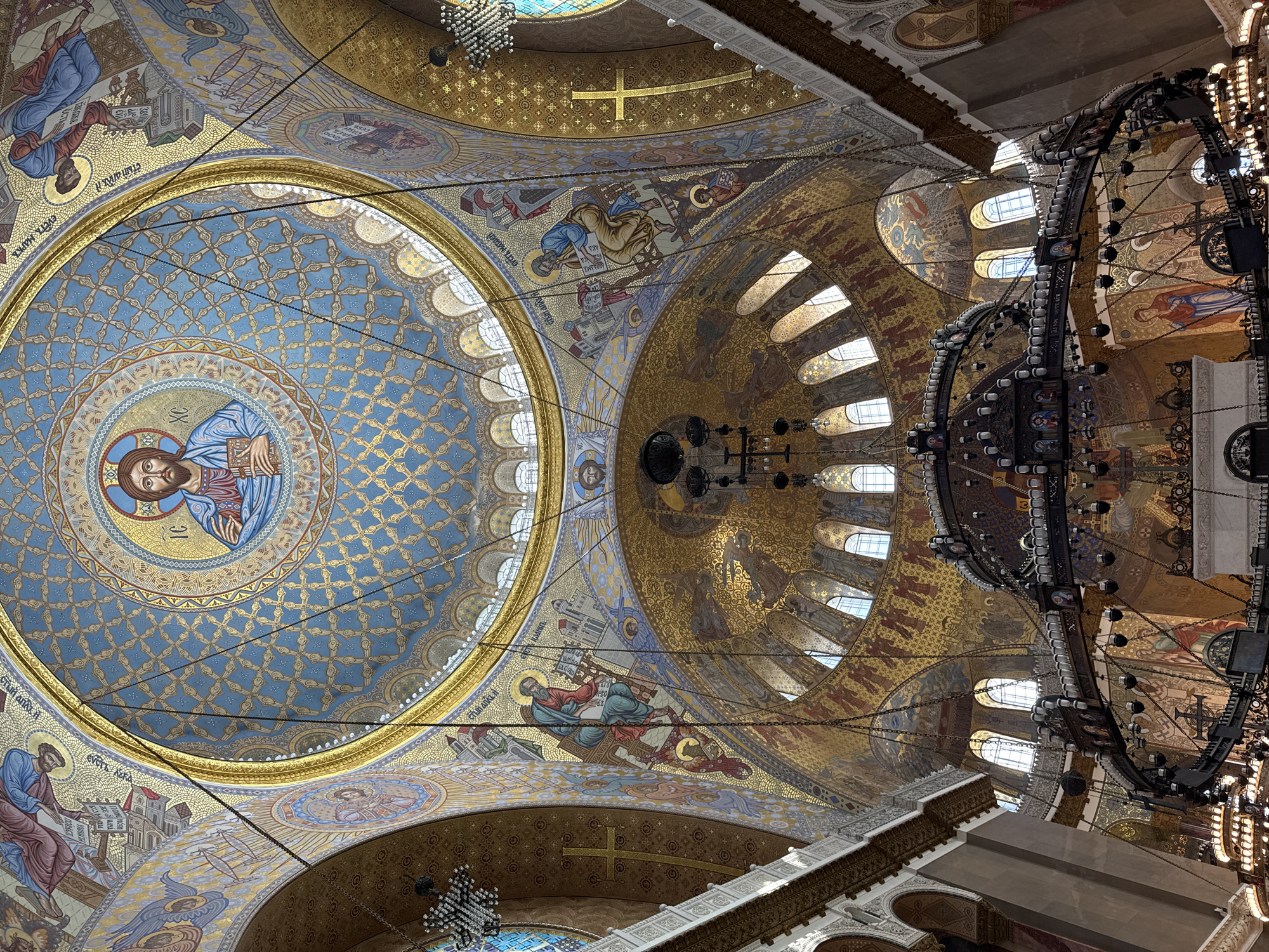 The image shows the interior of the Naval Cathedral of Saint Nicholas in Kronstadt, Russia. The view is upward towards the dome and ceiling of the cathedral. The dome is adorned with intricate mosaics depicting religious figures, primarily Jesus Christ in the center of the dome's highest point. Surrounding this central image are various scenes and figures from biblical narratives, rendered in gold and blue hues. The ceiling is also covered in mosaics and features arched windows that allow natural light to filter in. Large, ornate chandeliers hang from the ceiling, illuminated by numerous light bulbs. The architecture exhibits classical and Byzantine influences, with elaborate decorative elements, including gilded crosses and architectural mouldings. The overall impression is one of grandeur and spiritual solemnity.