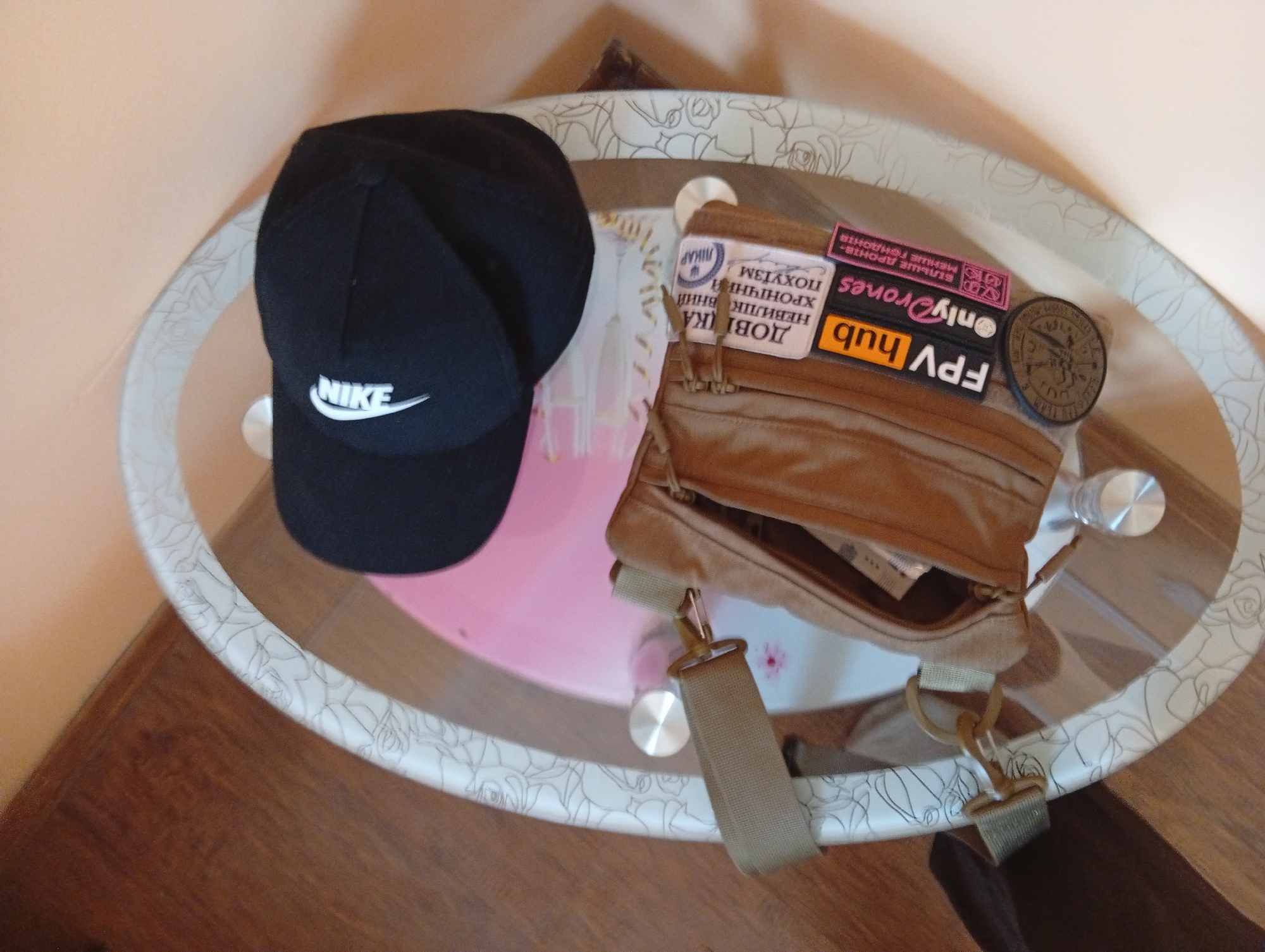 A black Nike baseball cap and a beige tactical bag with several patches are sitting on a small, oval-shaped table. The patches on the bag read "FPV hub", "OnlyDrones", and have text in Ukrainian. The table has a white floral pattern on its edge and metal legs. The table is situated on a dark wood floor. The location may be a private residence in Poltava, Ukraine, based on the Ukrainian text.