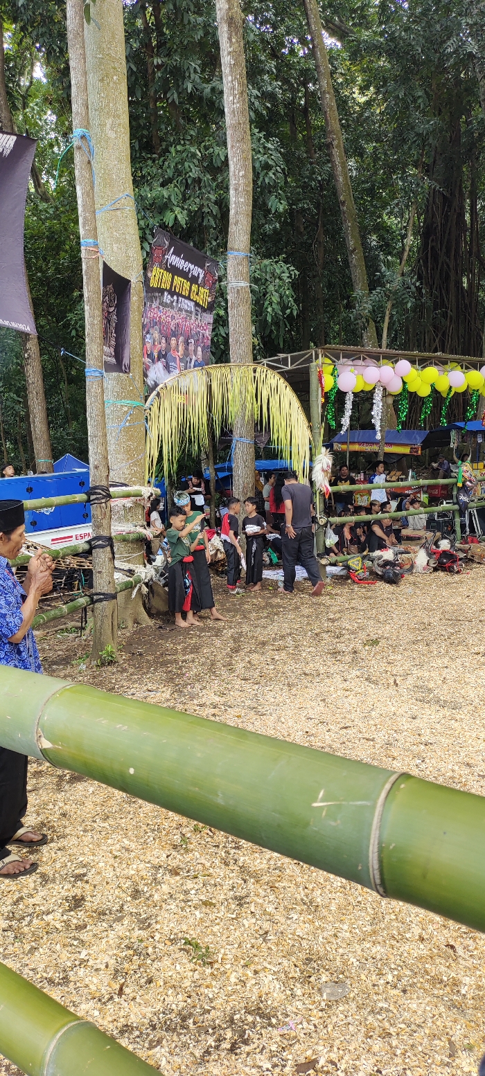 The scene is a gathering of people in a wooded area in Kecamatan Lawang, Indonesia, located at latitude: -7.844, longitude: 112.688. The group is sitting on the ground, which is covered in wood chips. There are several trees in the background, and a sign with the word "Anniversary" is hanging on one of them. The people are dressed in various casual clothes. Several people are sitting on a bench, while others are standing around. There are several objects scattered around the area, including a blue tarp, a sign that says "ESPA," and a variety of other items. There is also a large piece of bamboo on the ground in the foreground. The area is well-lit and there are no signs of any conflict or tension.