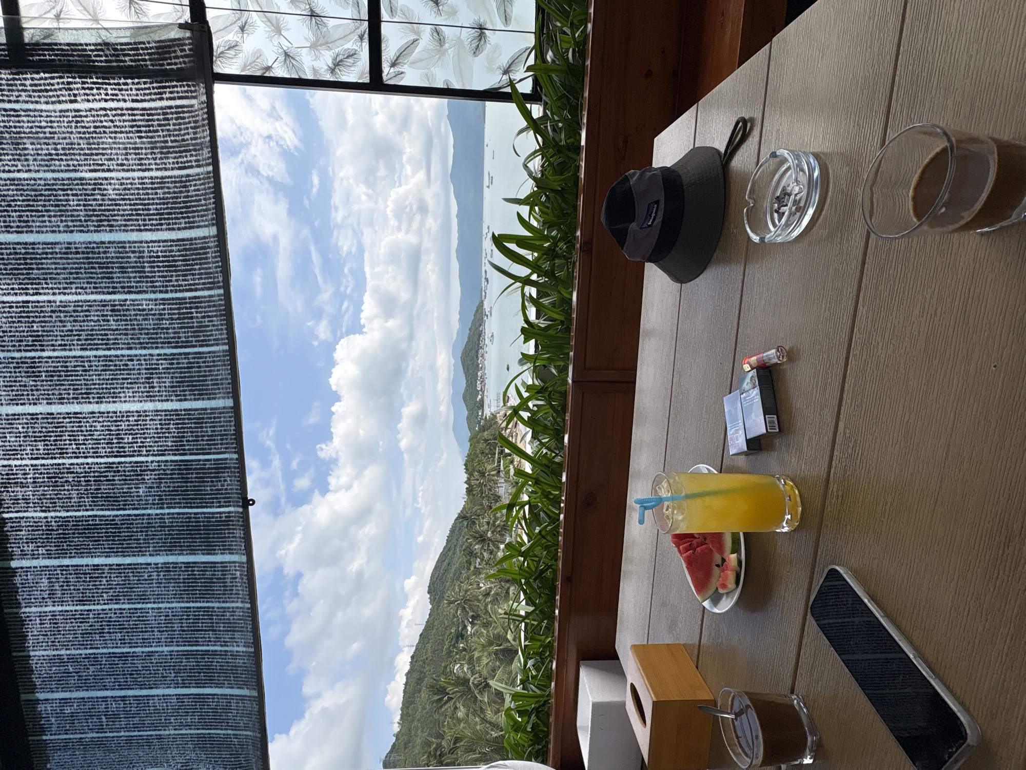 The image shows a table setting at a cafe or restaurant overlooking the city of Bac Nha Trang Ward, Vietnam. The view outside includes a coastline, a body of water, and a partially cloudy sky.
On the table are several items: a hat, an ashtray, a glass of coffee, a lighter, a pack of cigarettes, a glass of juice with a straw, slices of watermelon on a plate, a tissue box, another glass of coffee, and a smartphone. The table is made of a light brown material and is located within an establishment that has a partially shaded area with a patterned glass enclosure. Green foliage is visible between the table and the outside view.