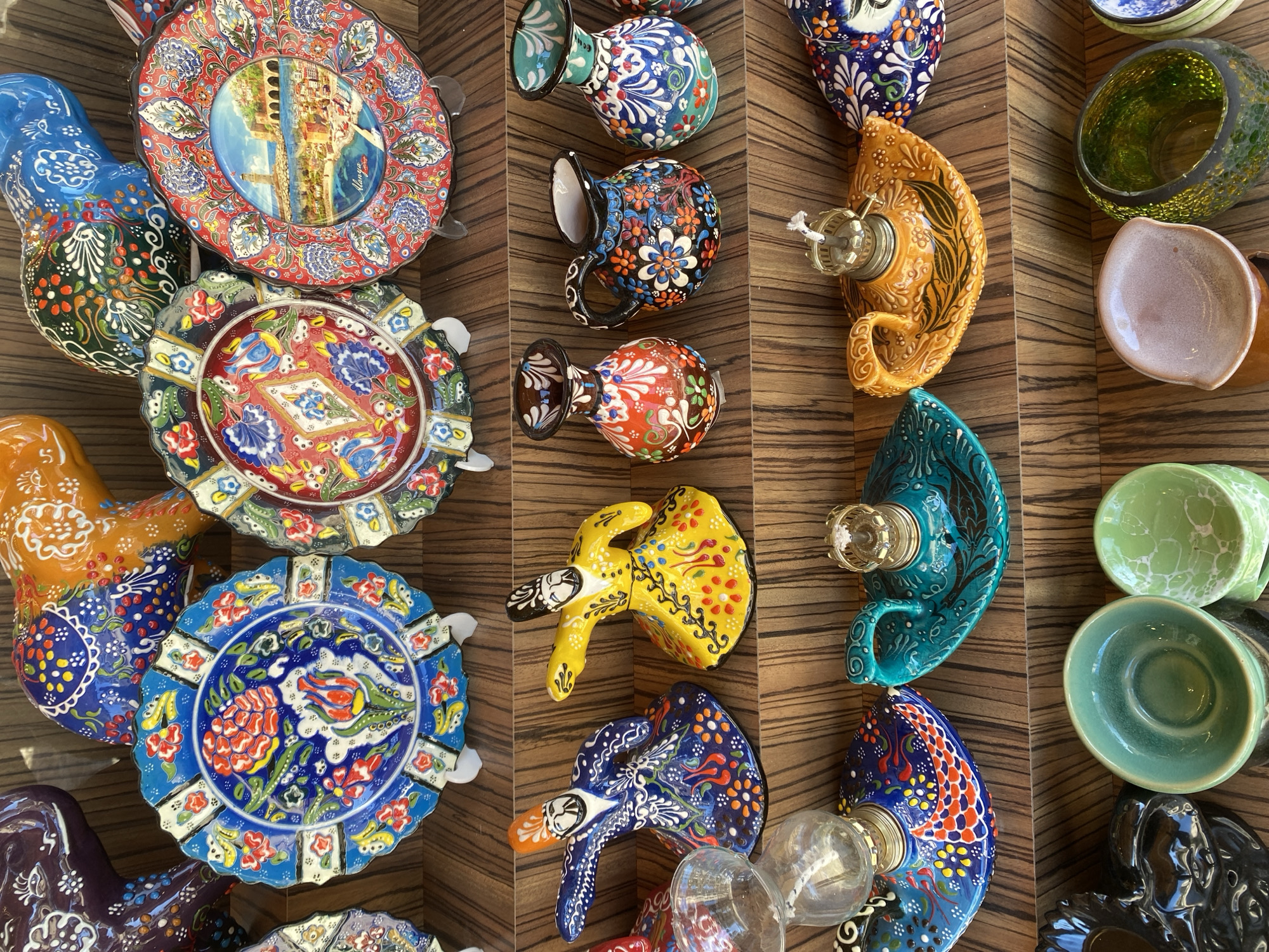 The image depicts a collection of colorful, hand-painted ceramic objects for sale, likely as souvenirs, in Alanya, Turkey. The objects are arranged on a dark wood-grain surface, possibly a shelf or display table. The items include plates, jugs, lamps, and decorative figures, all featuring intricate, traditional Anatolian or Ottoman-inspired designs in bright blues, reds, greens, yellows, and oranges. The plates are round with scalloped edges. The jugs have curved handles. The lamps are shaped like crescent moons. The decorative figures resemble dancing dervishes. The items are oriented in different directions, indicating they were placed casually. The light seems to come from above.