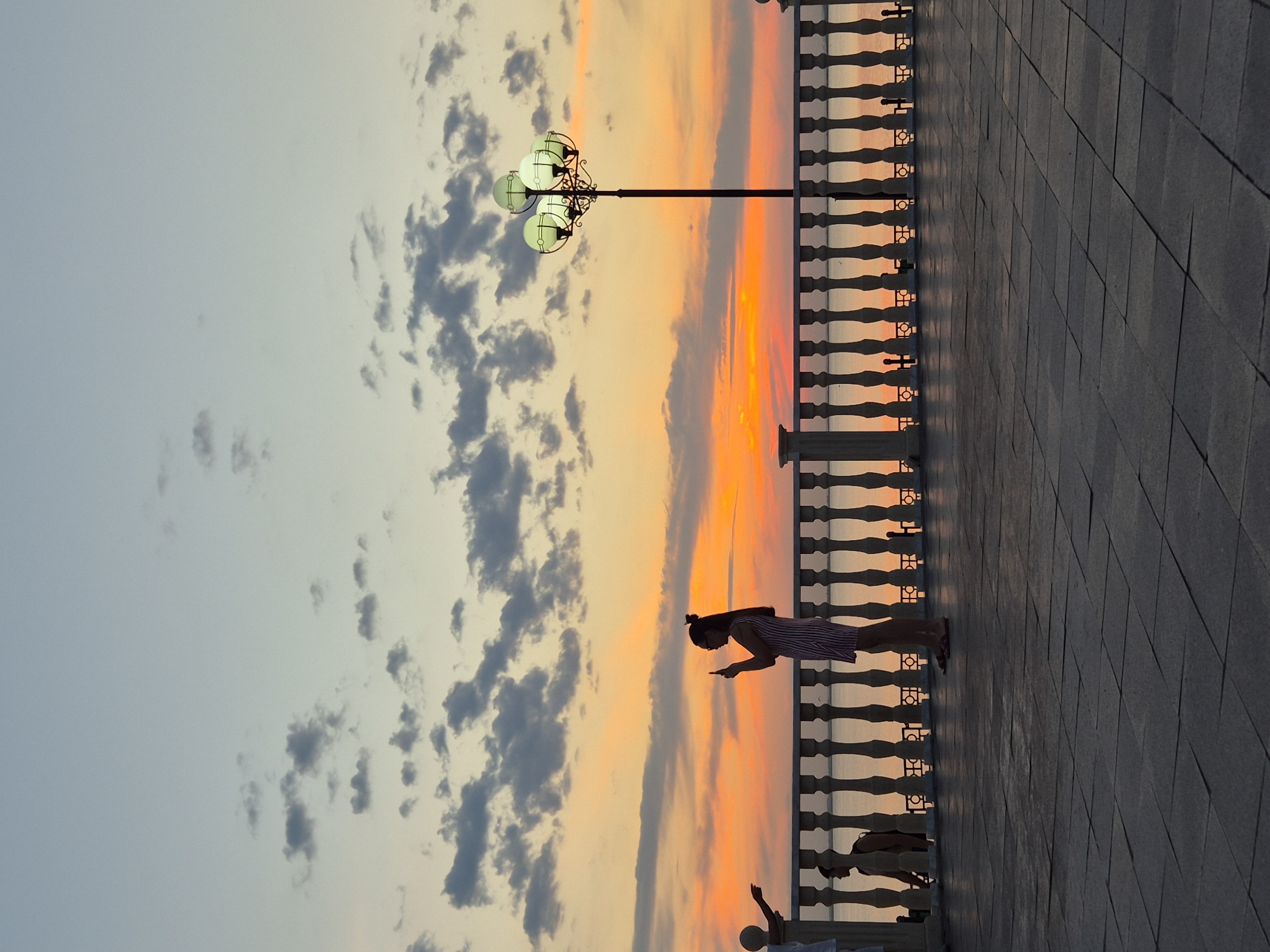 The image depicts a sunset scene along a paved walkway, possibly a promenade or boardwalk. The sky is ablaze with vibrant orange and yellow hues, indicative of the golden hour. Scattered clouds add texture to the sky.
A woman, silhouetted against the sunset, stands on the right side of the image, looking down at her phone. She is wearing a striped dress. To her left, a decorative lamppost with multiple globe-shaped lights stands prominently. A dark, ornate railing runs along the edge of the walkway, separating it from what appears to be a body of water or a lower level beyond. The ground is paved with large, dark tiles. In the lower portion of the image, a partial silhouette of another person can be seen. The overall mood is serene and peaceful, capturing the beauty of a sunset. The location is Kokshetau, Kazakhstan.