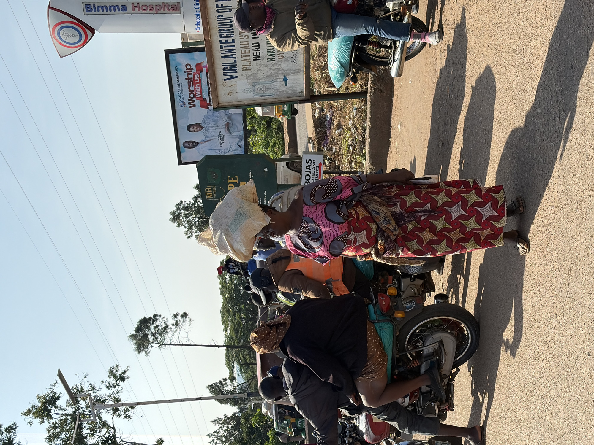 The image depicts a street scene in Gura Top, Nigeria, under a clear sky. A woman in the foreground, wearing a pink and red patterned dress and a headwrap, carries a large sack of goods on her head. She is walking on a paved road where other people and vehicles are present. To her left, two individuals are seated on a motorcycle; the person in front is wearing a grey jacket, and the person behind is wearing a black jacket and a patterned headscarf. Further behind, a man in an orange vest is also on a motorcycle. In the background, a sign for "Bimma Hospital" is visible, along with advertisements for "Omega Fire Ministries" and "Vigilante Group of Plateau State". Trees and power lines are also part of the environment. The overall atmosphere suggests a bustling urban or semi-urban environment.
