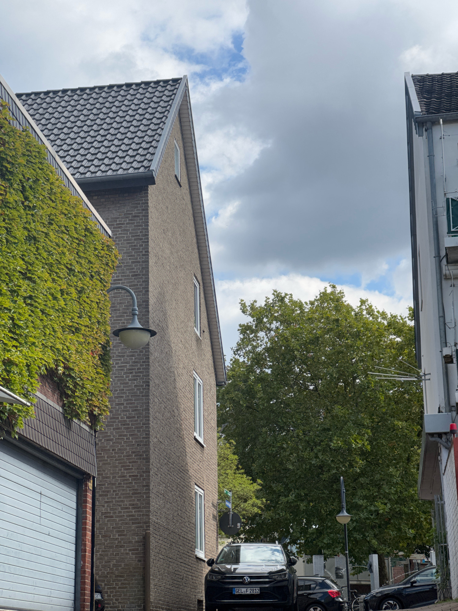 The image shows a street scene in the city of Cleves, Germany. On the left is a building with a garage door and a wall covered in green ivy. Adjacent to this is a brick building with a sloping grey tiled roof and several white-framed windows. A vintage-style outdoor lamp is mounted on the exterior wall of this building. In the background, a large deciduous tree with lush green foliage dominates the view. To the right, another building with a white facade and a sloping grey tiled roof is visible. Several cars are parked on the street, including a black SUV with a license plate reading "GEL F 2812" in the foreground. The sky is partly cloudy with scattered white clouds against a blue backdrop.