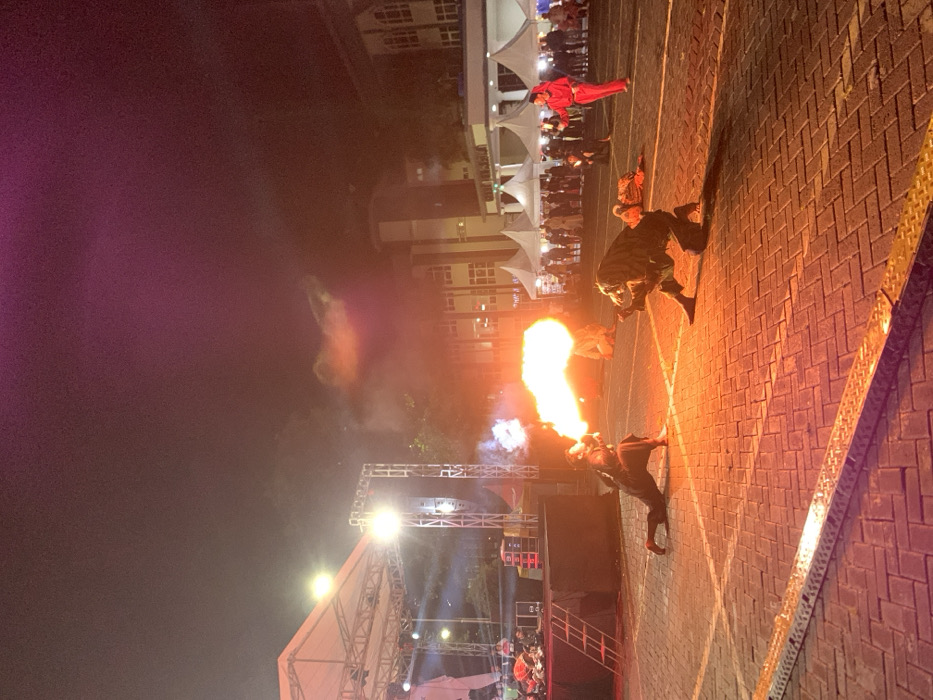The image shows a performance of fire breathing in the evening at an outdoor event in the city of Batu, Indonesia. The location is identified by the coordinates -7.867 latitude and 112.514 longitude. The performance takes place on a brick-paved square in front of a stage, surrounded by white tents. There are two fire breathers performing. The performer on the left is breathing fire, and the performer on the right is kneeling in a dramatic pose, with a large flame visible in the background. The performer on the left wears a black costume and a black hat. The performer on the right wears a dark green costume. In the background, there are people watching the performance, and several white tents, illuminated by white lights. There is a large stage to the left, partially visible, with a large white awning stretched over it. The sky is dark, and there is smoke in the air.
