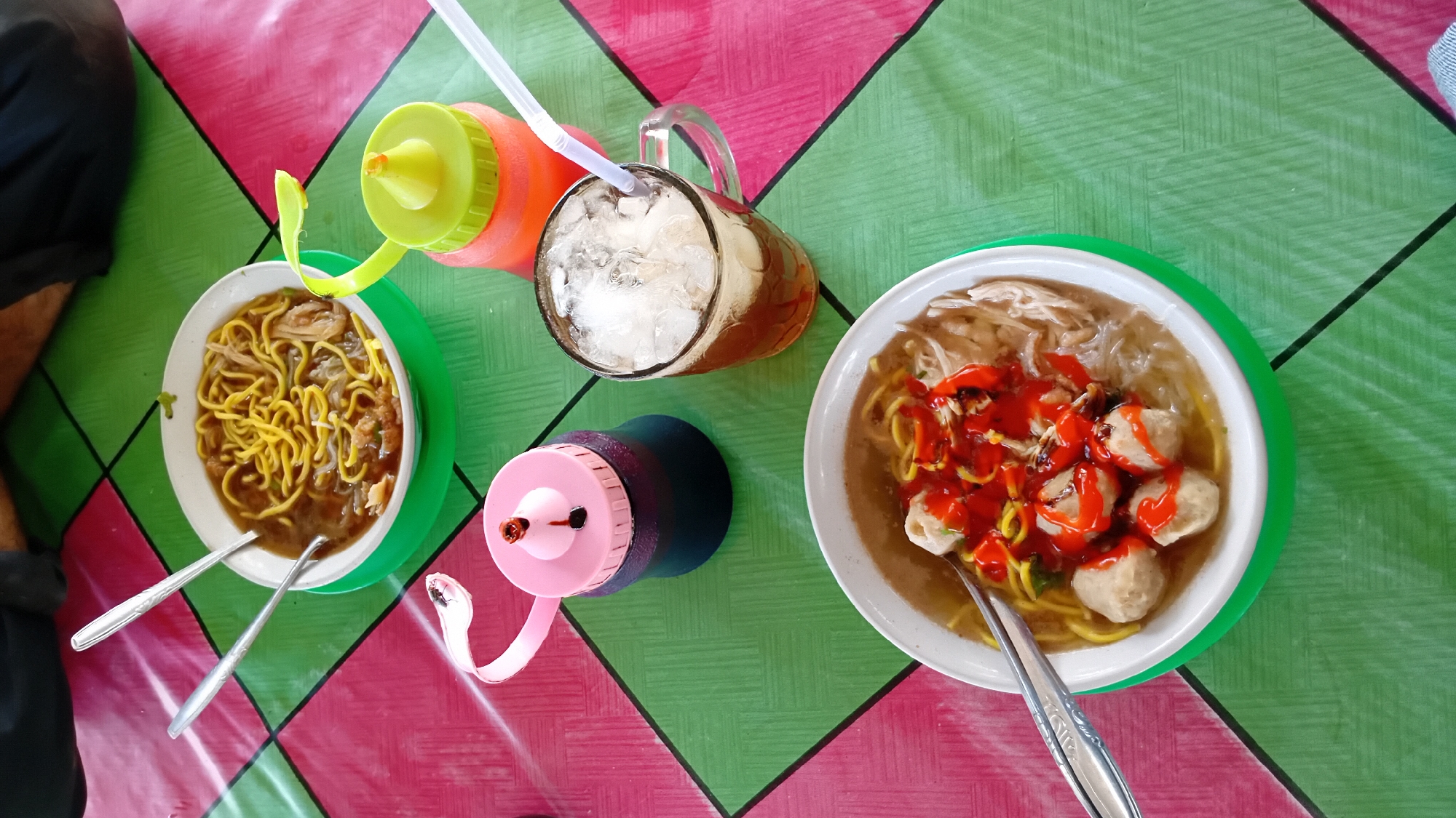 An overhead shot shows two bowls of noodles, a glass of iced tea, and two squeeze bottles on a green and pink checkered tablecloth. The bowl in the foreground contains noodles, meatballs, and a red sauce. The bowl in the background also contains noodles and broth. The glass of iced tea is filled with ice and a straw. One squeeze bottle is pink with a black lid, and the other is orange with a green lid and a green spout. The scene appears to be set in an outdoor dining area in Cotkumbang, Indonesia, possibly at a street food stall or a casual eatery. The lighting suggests it is daytime. No people are visible in the image.