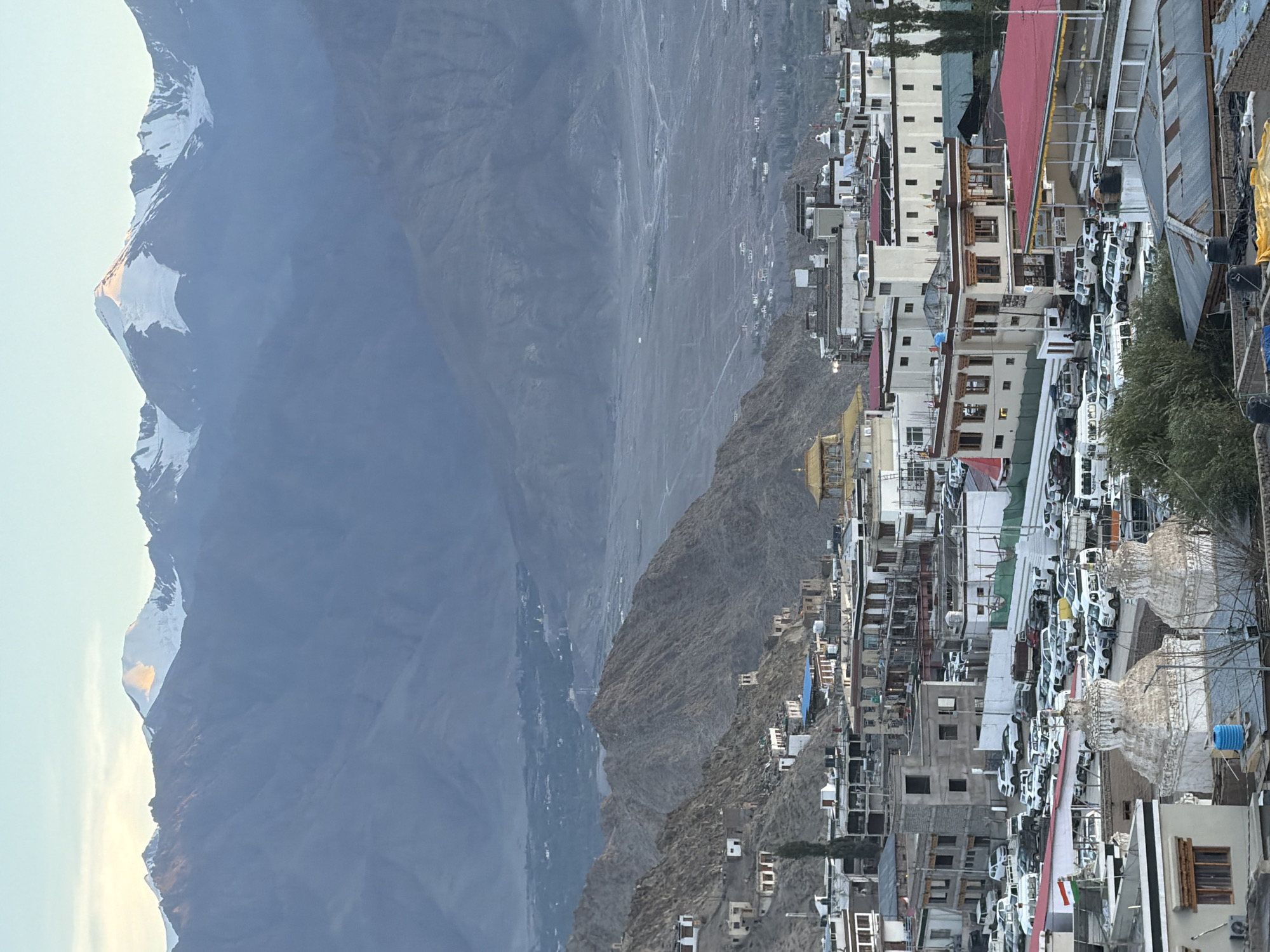 This is a high-angle, medium-shot image of a cityscape in Leh, India, with mountains in the background. The foreground features several buildings with flat roofs, some with intricate white stupa-like structures in front of them. A large parking lot filled with white SUVs and vans is visible in the middle ground. A few trees are scattered throughout the scene. In the far background, imposing snow-capped mountains rise against a hazy, pale blue sky. The overall impression is of a town nestled in a valley, surrounded by a rugged mountainous landscape. The lighting suggests it is either early morning or late afternoon, with soft sunlight illuminating the mountain peaks.