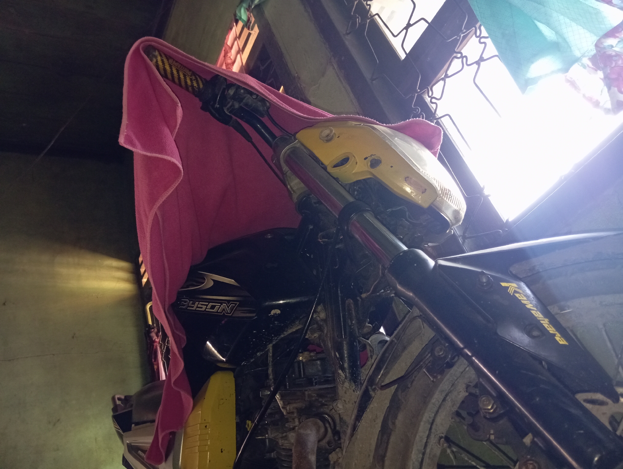 A low-angle, wide shot captures the front of a dirt-covered motorcycle in Rejosari, Indonesia. A bright pink cloth is draped over the handlebars and front fairing, obscuring part of the yellow-painted headlight assembly. The motorcycle's front wheel, with a visible tire, is in the foreground. The background features a window with a metal grate and sunlight filtering through, along with some draped fabric, possibly clothes drying. The overall environment appears to be an outdoor or semi-outdoor space, possibly a yard or a workshop, with a green wall visible on the left. The motorcycle's make appears to be "BISON" as indicated on its side.