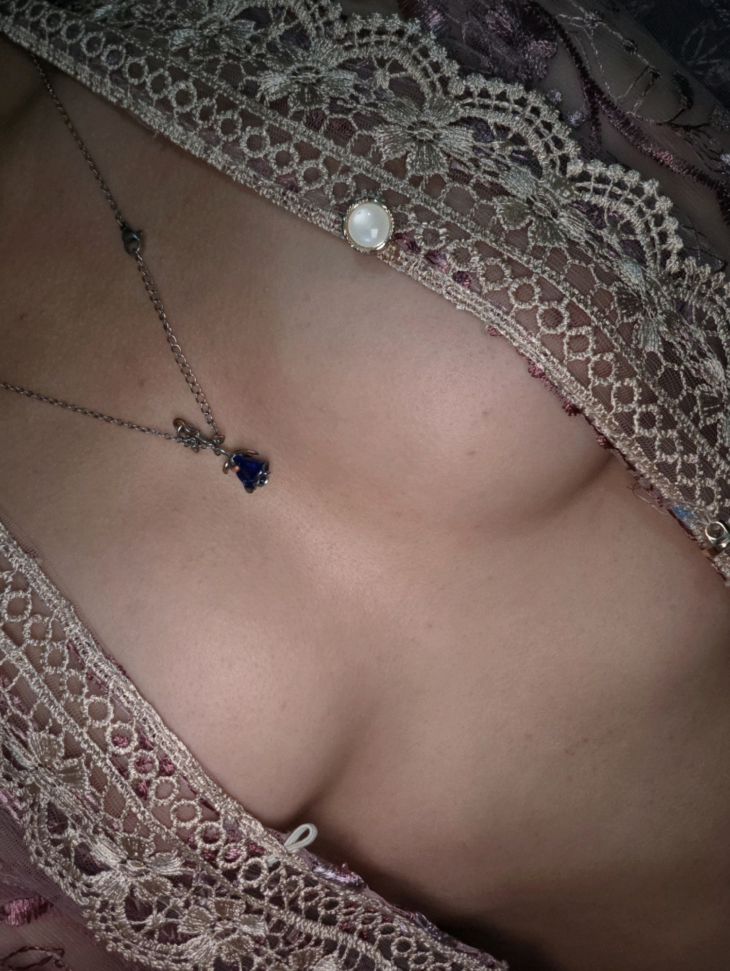 The image shows a close-up view of a person's chest and décolletage, adorned with a delicate, embroidered lace garment. A silver chain necklace with a small, dark blue gemstone pendant rests on the skin. A single pearl is visible on the lace near the collarbone. The lighting is soft, highlighting the textures of the lace and the skin. There is no discernible background or environmental context to suggest a specific location within Phan Thiết, Vietnam, or any other city.