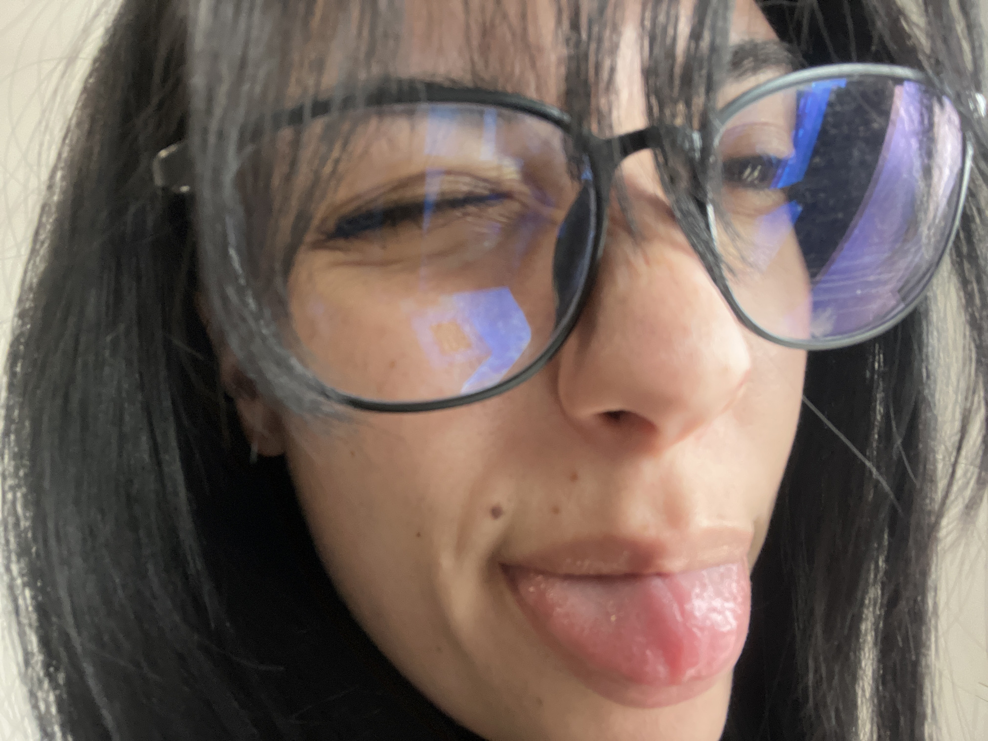 A close-up selfie shows a person with dark hair and bangs wearing glasses. They are sticking their tongue out. The reflection in their glasses shows a window. It is not possible to determine the location in Saedinenie, Bulgaria from the provided image.