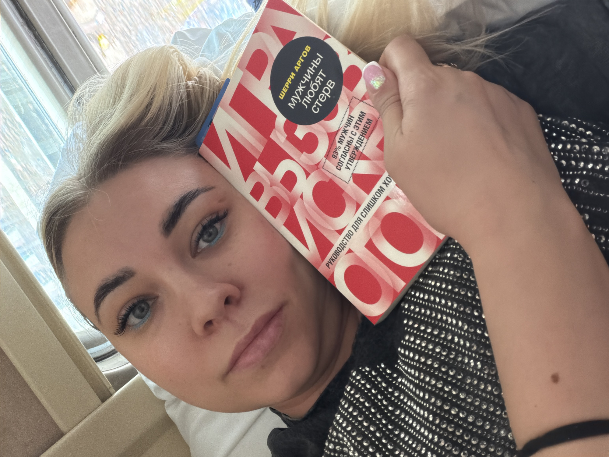 A woman lies in an interior setting, holding a book close to her face. She has fair skin, light blond hair in a bun, and light blue eyes with what appears to be blue eyeliner. She is wearing a black top with small, reflective embellishments. The book she holds is titled "ШЕРРИ АРГОВ МУЖЧИНЫ ЛЮБЯТ СТЕРВ" in Russian, which translates to "Sherry Argov Men Love Bitches" with other smaller text reading "93% мужчин согласны с этим утверждением," meaning "93% of men agree with this statement". The book's cover has a red and white design with bold, capitalized letters. The background shows a window with a blurry outdoor scene. The setting is assumed to be in Варламово, Russia, as per the user's prompt.