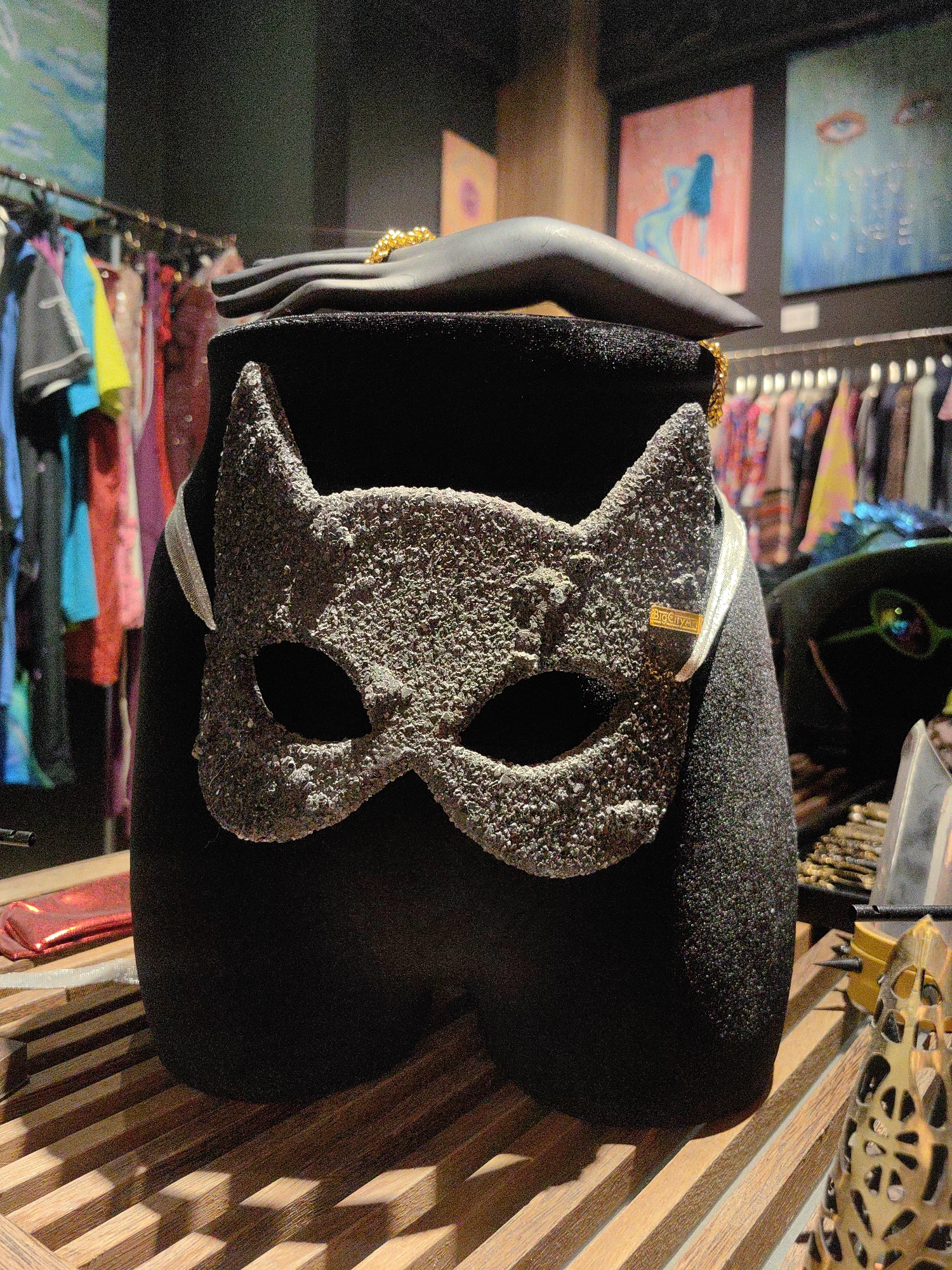 In Baan Mae Nam, Thailand, a black mannequin leg wears a textured gray cat mask. The mask has two eye holes and pointed ears. A gold chain is draped around the mannequin's neck and over its shoulder. Behind the mannequin, racks of colorful clothing are visible. The mannequin stands on a wooden surface.