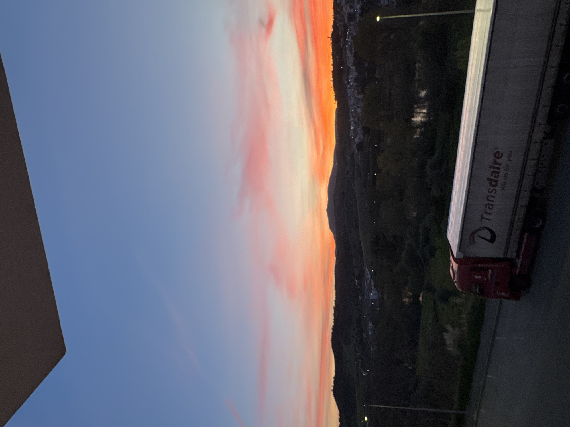 The image depicts a truck with the logo "Transdaire - We do for you" parked on a road in Mirandela, Portugal. Behind the truck, there's a cityscape bordering what appears to be a forested or mountainous area, illuminated by streetlights. The sky is a gradient of blue and orange, suggesting either sunrise or sunset. The perspective suggests the photo was taken from an elevated position, potentially from a building or higher ground nearby. The scene is captured at dusk or dawn given the lighting conditions. A corner of a building appears to be visible in the left corner of the image.