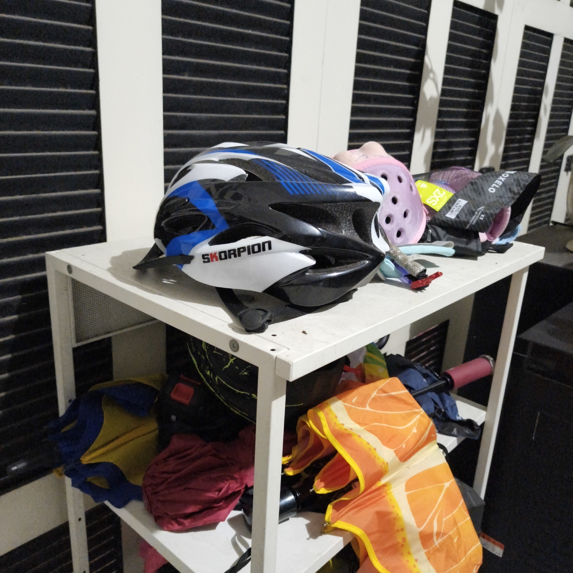 This image captures a cluttered shelf within an indoor space, possibly a storage room or garage, located in South Jakarta, Indonesia. The shelf is white and appears to be made of metal and possibly particle board. On the top shelf, a black, white, and blue bicycle helmet with "SKORPION" written on it is prominently displayed. To the right of the helmet are several other items including a pink Croc-like shoe, a purple hat or beanie, and a black bag with white lettering that reads "OXELO".
The shelf below contains a collection of items, including what appears to be folded umbrellas in blue and yellow, a red folded item, and an orange and yellow patterned fabric, possibly another umbrella or bag. A dark object with a pink handle, potentially a bicycle component or tool, is also visible on this lower shelf.
The background consists of vertical black slats or blinds against a white wall, casting faint shadows. The overall impression is one of disarray and storage of various personal belongings. There are no people visible in the image. The lighting is dim, suggesting an indoor environment.