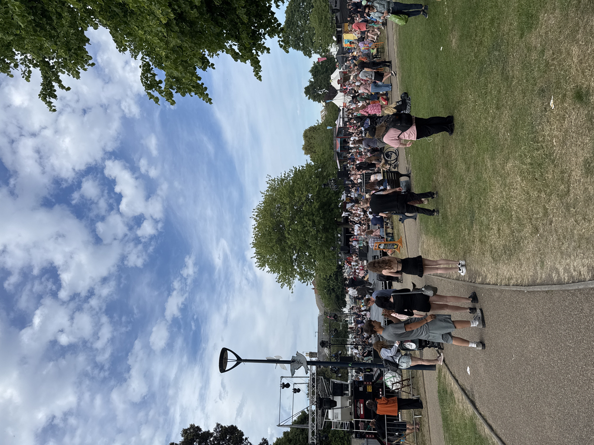 This photograph captures a daytime outdoor event in Great Yarmouth, United Kingdom. A crowd of people, appearing diverse in age and attire, are gathered on a grassy area. Some are seated on chairs or on the ground, while others are standing and walking. A stage is visible in the lower portion of the image, along with sound equipment and scaffolding, suggesting a performance or event. A prominent lamppost stands near the stage, featuring a bird sculpture at its top. The sky is partially cloudy with areas of blue. Trees are visible at the periphery of the frame. A paved path runs along the bottom of the frame, with several people standing and walking on it.