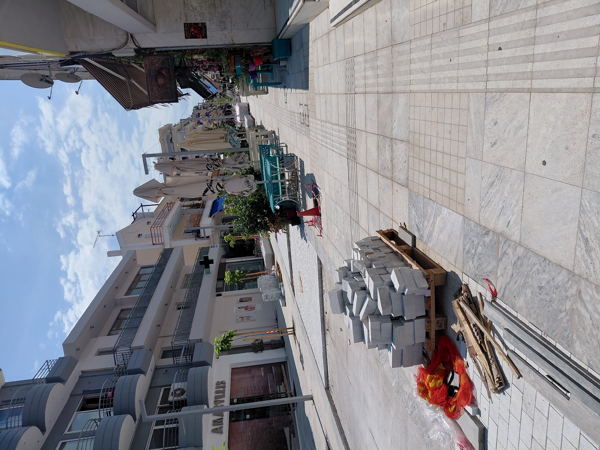 This is a street scene in Aigio, Greece, on a bright, sunny day with scattered clouds. The image captures a street undergoing renovation or construction. In the foreground, a stack of light-colored, rectangular paving stones rests on a wooden pallet. Adjacent to the pallet are scattered pieces of wood, some plastic sheeting, and a bright orange mesh bag.
The street is paved with large, light-colored marble tiles, some of which have a textured, tactile paving pattern for the visually impaired. The sidewalks are also paved with similar tiles. On the left side of the street, a building with multiple balconies, painted in gray and white, is visible. The ground floor of this building appears to be a shop with a closed metal shutter and a sign that reads "AMARYLLIS."
Further down the street, on the right side, are outdoor seating areas with tables and chairs, suggesting a cafe or restaurant. Some tables are covered with umbrellas. The buildings lining the street are multi-story structures, typical of urban environments. There are no people visible in the immediate foreground or midground. The overall impression is one of ongoing work and preparation for reopening or refurbishment.