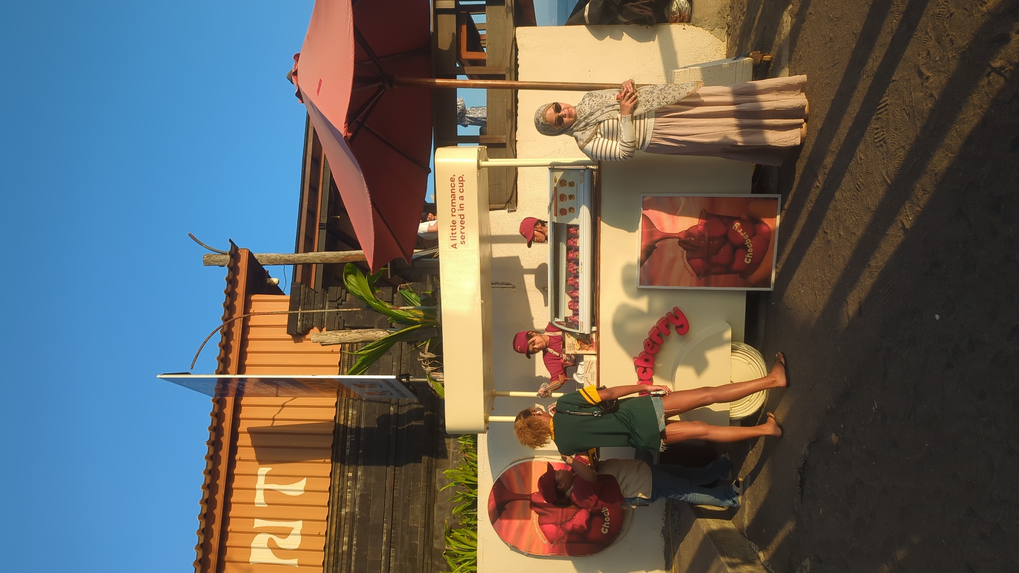 In the city of North Kuta, Indonesia, a curbside food stall is depicted under a clear blue sky. The stall, painted in a light cream color, features a display case filled with what appear to be chocolates or candies. A red umbrella provides shade over a portion of the stall. Three individuals are visible around the stall. Two employees, wearing matching maroon caps and shirts, are behind the counter. One is serving a customer, a young woman wearing a green and yellow striped shirt, denim shorts, and sandals, who stands facing the counter. Another employee is partially visible behind the counter, to the right. A woman wearing a hijab and a long skirt stands to the right of the stall, holding a drink. In front of the stall, a framed advertisement displays a close-up of chocolate being poured over strawberries in a cup, with the text "Choccherry" visible. The ground is covered in dark sand, with strong shadows cast by the stall and umbrella, indicating late afternoon sunlight. Behind the stall, a building with an orange corrugated metal roof and dark stone accents is visible.