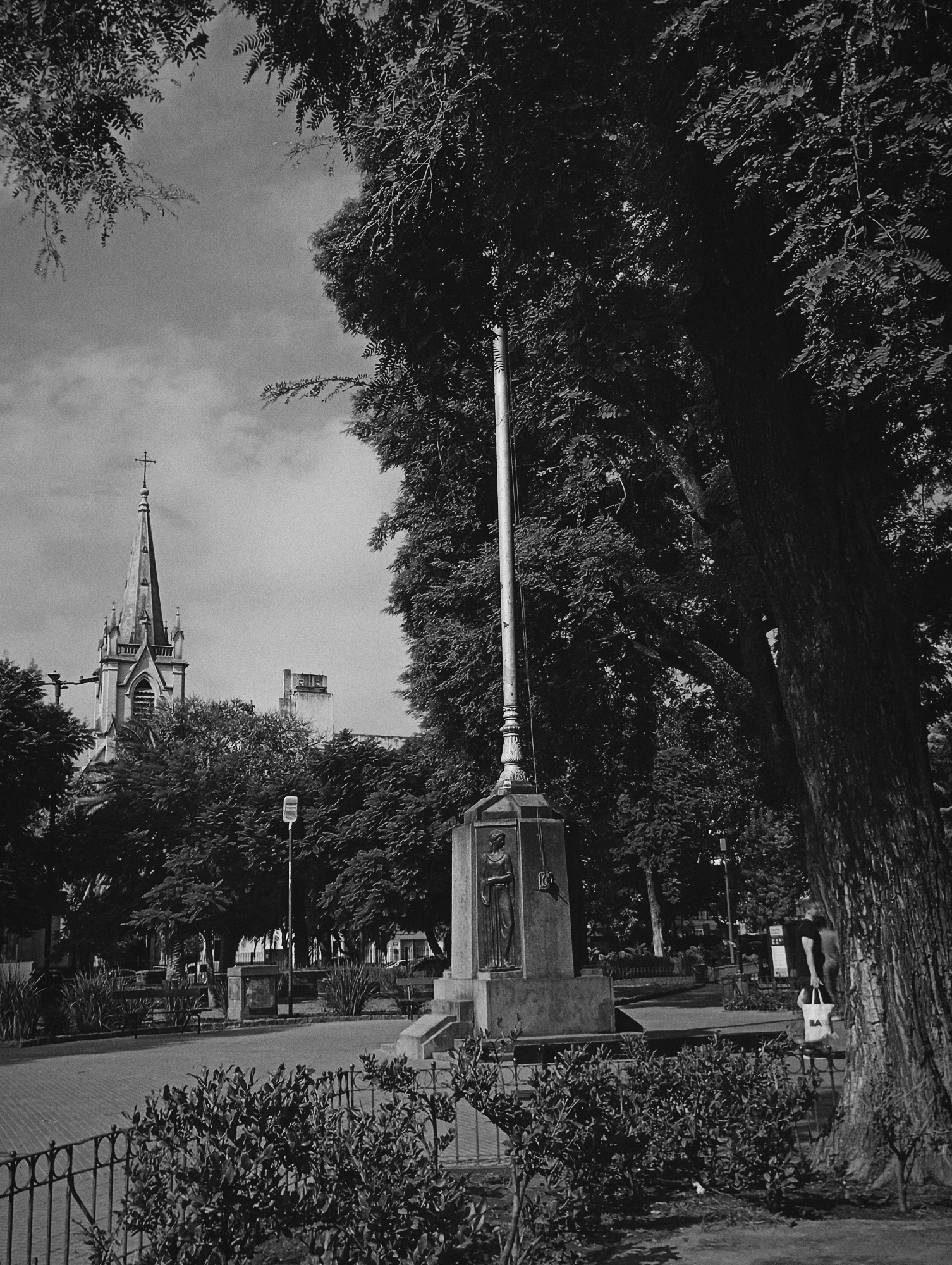 The black and white image shows a section of a park in Buenos Aires, Argentina. In the left background, a church steeple rises above the trees, its cross distinct against the sky. The park features a stone monument with a pole extending upwards, positioned to the right of the church. The monument has a relief sculpture of a person on its front face. A large tree dominates the right side of the frame, its dark foliage casting shadows on the scene. In the foreground, a wrought-iron fence surrounds a small patch of bushes. A pedestrian with a shopping bag can be seen to the right, walking past the monument. The park appears to be sparsely populated, with benches visible in the background.