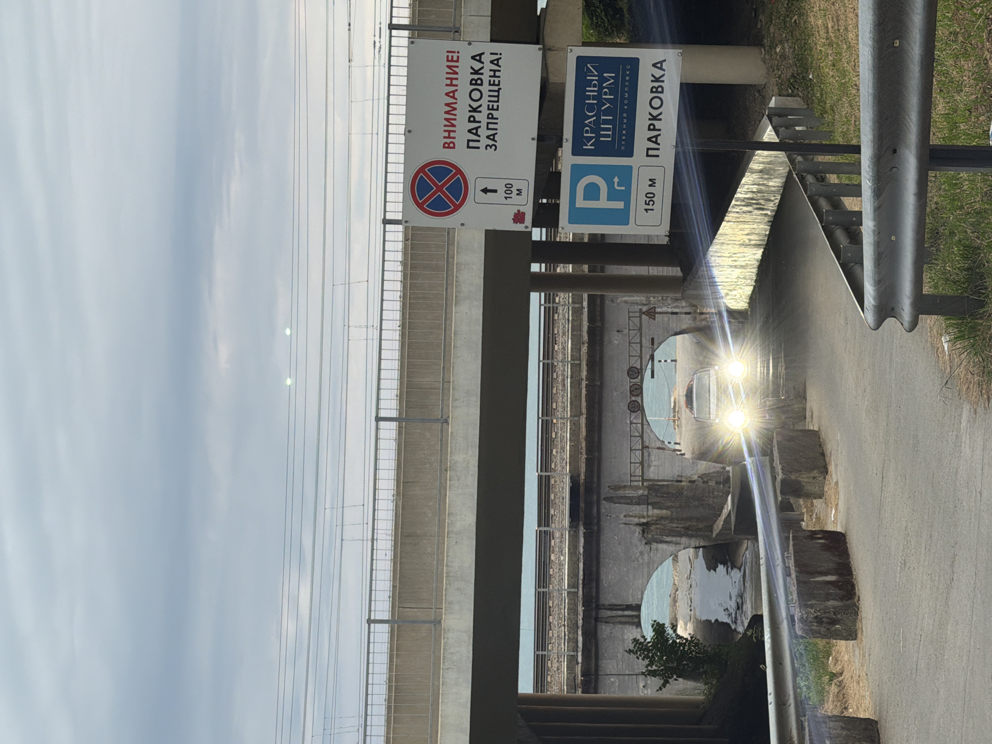 The image captures an outdoor scene with a road curving towards an underpass. A car with its headlights on is approaching the underpass, causing glare. Several signs are visible on the right side of the road. The top sign, rectangular and white, contains a circular red sign with a blue cross and text in Russian indicating "WARNING! PARKING IS PROHIBITED!". Below it, a smaller sign shows an arrow pointing upwards and "100 M". The second sign, also rectangular and white, has a blue "P" symbol for parking, a curved arrow, and the text "KRASNY SHUTRM", followed by "BEACH COMPLEX" and "PARKING 150 M". The environment includes concrete barriers, grass, and foliage. In the background, a concrete overpass with railings is visible, with train tracks running on it. Underneath this, another bridge structure with arched openings is present. The sky is overcast with grey clouds. The location appears to be in Sochi, Russia, given the Russian text on the signs.
