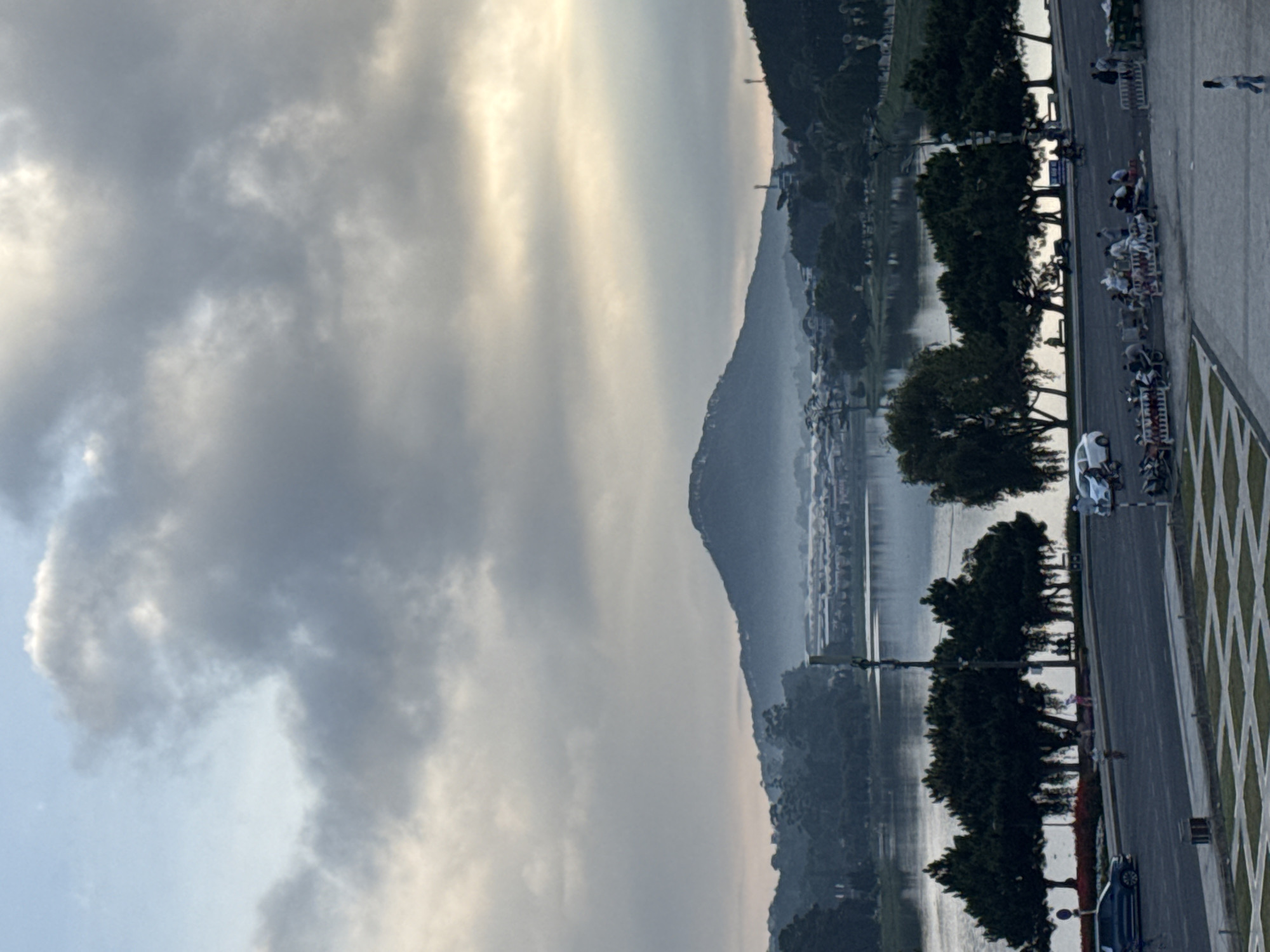 The image captures a landscape scene in Da Lat, Vietnam. The sky is filled with clouds and sunlight. In the distance, a mountain range is visible, shrouded in mist. A body of water reflects the sky. Alongside the water is a road lined with trees. People are present on the road, some on foot. Several parked vehicles are noticeable on the side of the road. A patterned walkway borders the road.