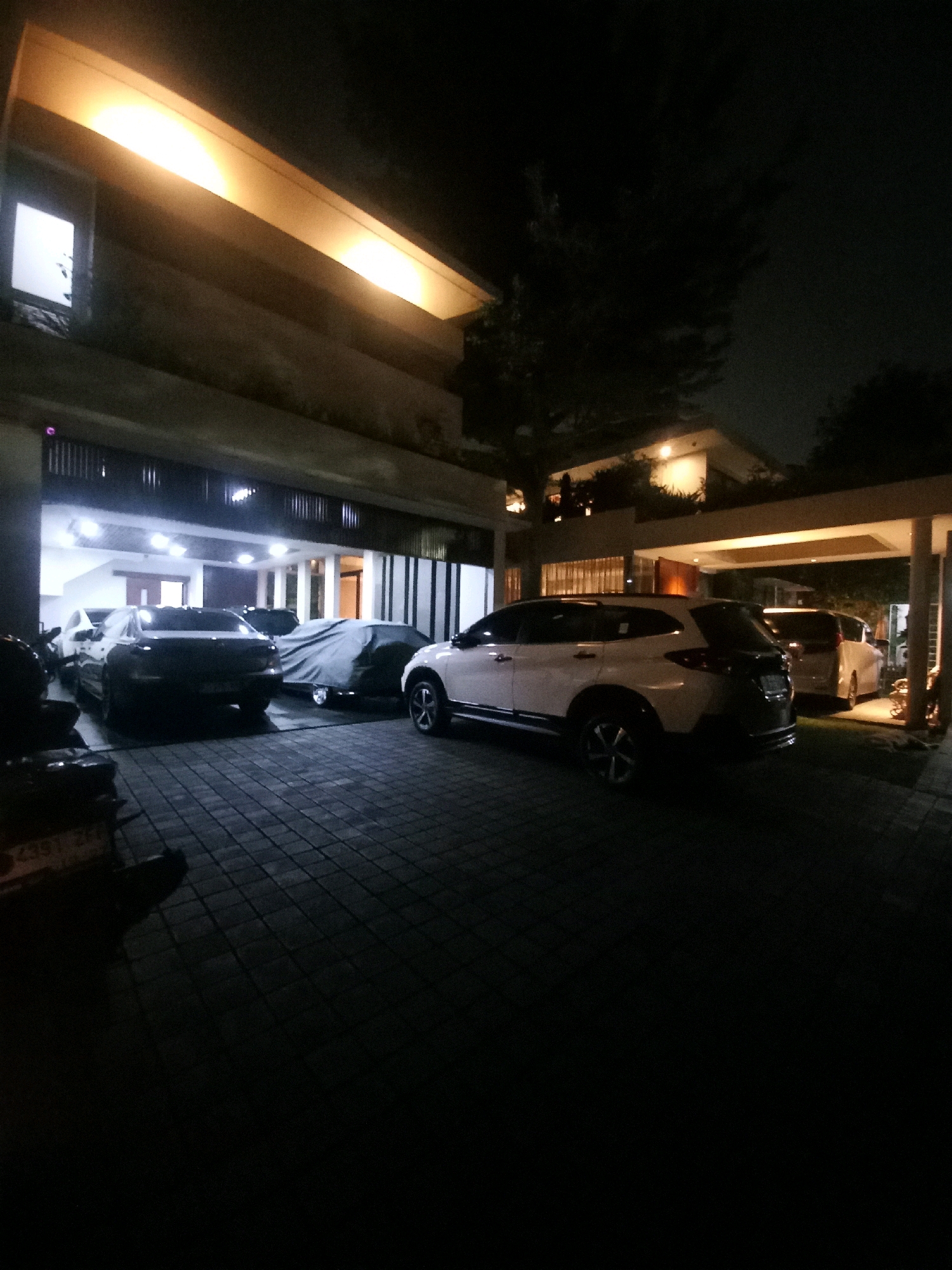 A nighttime exterior shot captures a modern residence in South Jakarta, Indonesia, characterized by its sleek architecture and integrated garage. The scene is illuminated by warm, recessed lighting that highlights the building's overhangs and the driveway.
In the foreground, a white SUV is parked. The driveway is paved with dark, square interlocking bricks, creating a textured surface that reflects the ambient light. To the left, several other vehicles are partially visible. One appears to be a dark sedan, and another is covered by a gray tarp. Further back, in the open garage area, at least two other vehicles are parked, their forms illuminated by interior lighting.
The environment suggests a well-maintained private property, with the lush darkness of the night sky overhead. The overall impression is one of a secure and well-lit residential space in a metropolitan area.