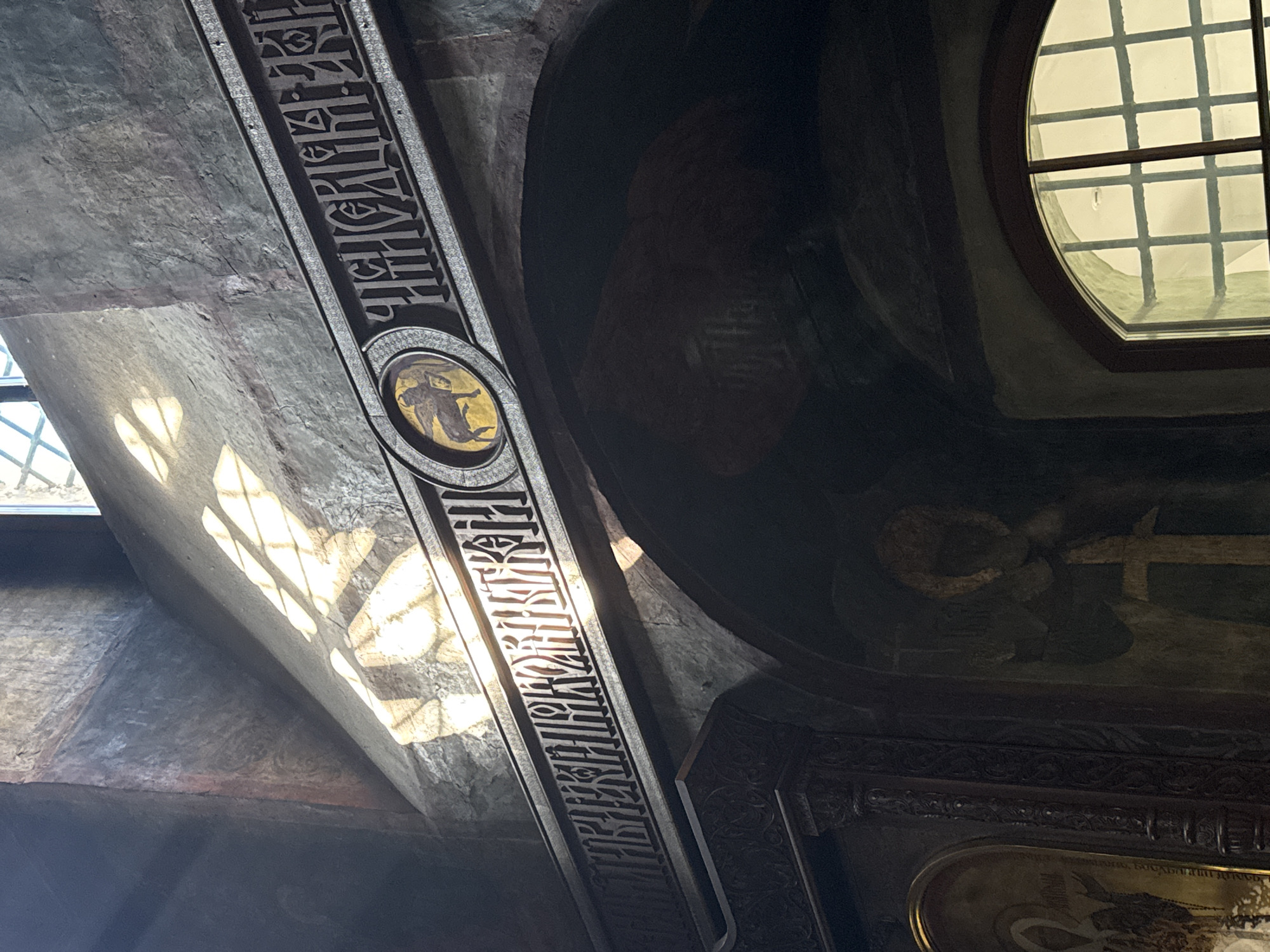 The image depicts the interior of a dimly lit religious building, likely a church, with sunlight streaming in from a high window. The architectural elements suggest a historical setting, possibly in Moscow, Russia, given the prevalent ornate carvings and religious iconography.
In the upper portion of the image, a textured wall is illuminated by sunlight creating distinct patterns of light and shadow. A horizontal band with intricate Cyrillic inscriptions runs across the wall, bordering a circular medallion containing a silhouette of a winged figure, possibly an angel or saint, on a yellow background. Below this, an arched opening reveals a darker interior space, where faint religious paintings or frescoes can be observed on the curved ceiling. To the left, a carved wooden pillar is visible, part of a larger ornate structure. Further to the left, a framed icon with a religious figure is partially visible. On the right side of the lower arch, a window with a grid pattern is illuminated from the outside, casting light into the interior. The overall impression is one of ancient religious artistry and solemnity, enhanced by the play of light and shadow.