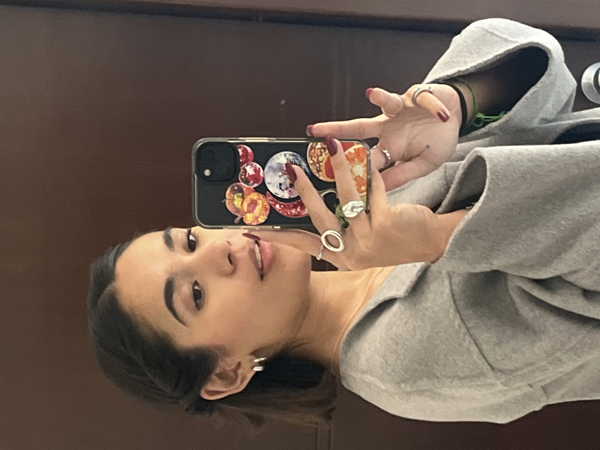 A young woman is captured in a mirror selfie, posing indoors in what appears to be San Bernardino Tlaxcalancingo, Mexico. She is positioned with her head tilted, looking directly towards her reflection in the phone she holds in her right hand. She has dark hair, styled down, and dark eyes. Her lips are slightly parted, and her expression is calm.
She is wearing a light grey hooded sweatshirt or jacket, a thin gold chain necklace, and small silver hoop earrings. Her fingers are adorned with several rings, including simple bands, a larger abstract silver ring, and one with a circular motif. A thin green bracelet is visible on her left wrist, and her fingernails are painted a dark red or burgundy.
The black smartphone she holds features a distinct decorative case adorned with various circular and oval graphics. These graphics depict what appear to be fruits, such as a sliced orange or grapefruit, apples or peaches, and clusters of red berries, alongside what could be a stylized moon or planet. The background behind her is a solid, dark brown surface, likely a wall or furniture, contributing to the indoor setting. There is no visible text in the image.