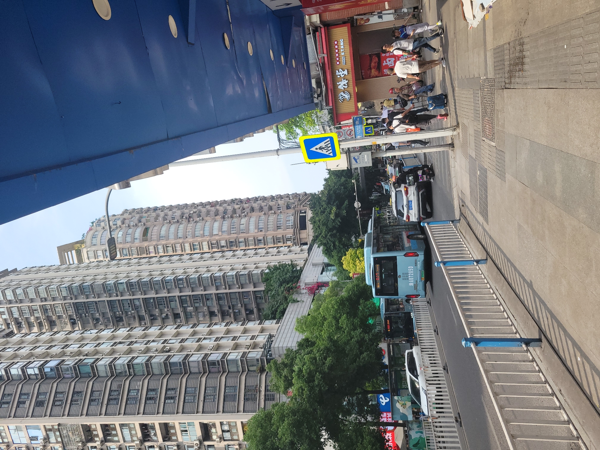 The image depicts a street scene in Shapingba District, China. On the right side of the image, a pedestrian crosswalk is visible, with people crossing the street. A blue crosswalk sign is posted above the crosswalk. A bus and cars are driving along the street. Tall apartment buildings line the left side of the image. A blue overhead structure is visible above the street.