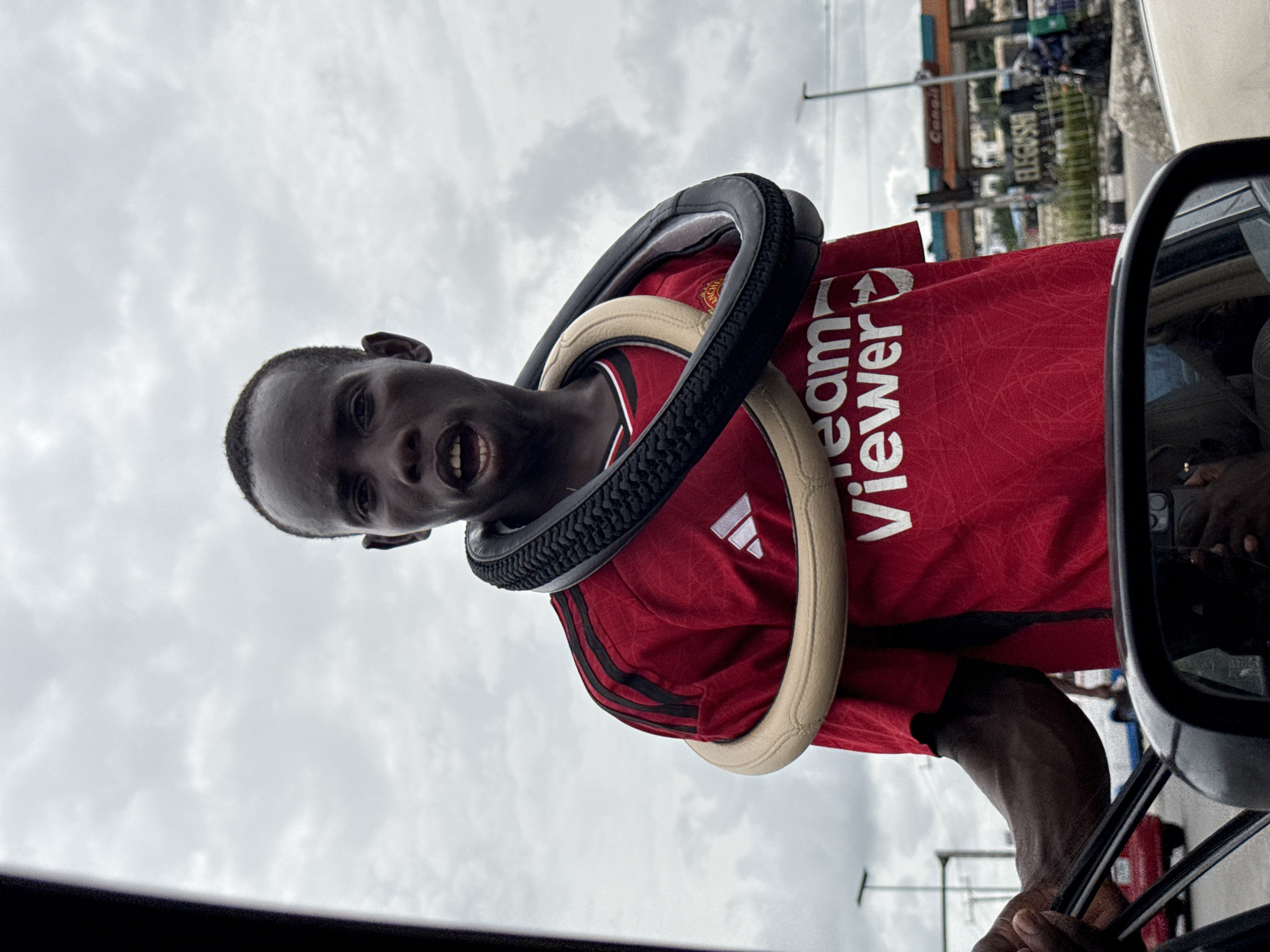 A young Black man, likely a street vendor, is captured looking directly into the camera from outside a car. Two steering wheel covers, one black and textured, the other cream-colored, are draped around his neck, suggesting items he is selling. He is wearing a red short-sleeved Manchester United football jersey, featuring the white "TeamViewer" sponsor logo prominently on the chest, along with the Adidas logo and the club crest. His mouth is slightly open, and he maintains direct eye contact with the viewer.
The setting is an urban or semi-urban street in Alaguntan, Nigeria, under an overcast sky, indicating daytime. In the background, several buildings and structures are visible, including one with "Conoil" (a gas station brand) and another with "ELLEGUSHU" written on it. A power line structure is also visible against the cloudy sky. Part of the car's side mirror is seen on the right, and a glimpse of the car's interior, possibly the dashboard or a hand, is visible at the bottom right. The image captures a candid moment of street commerce and interaction.
