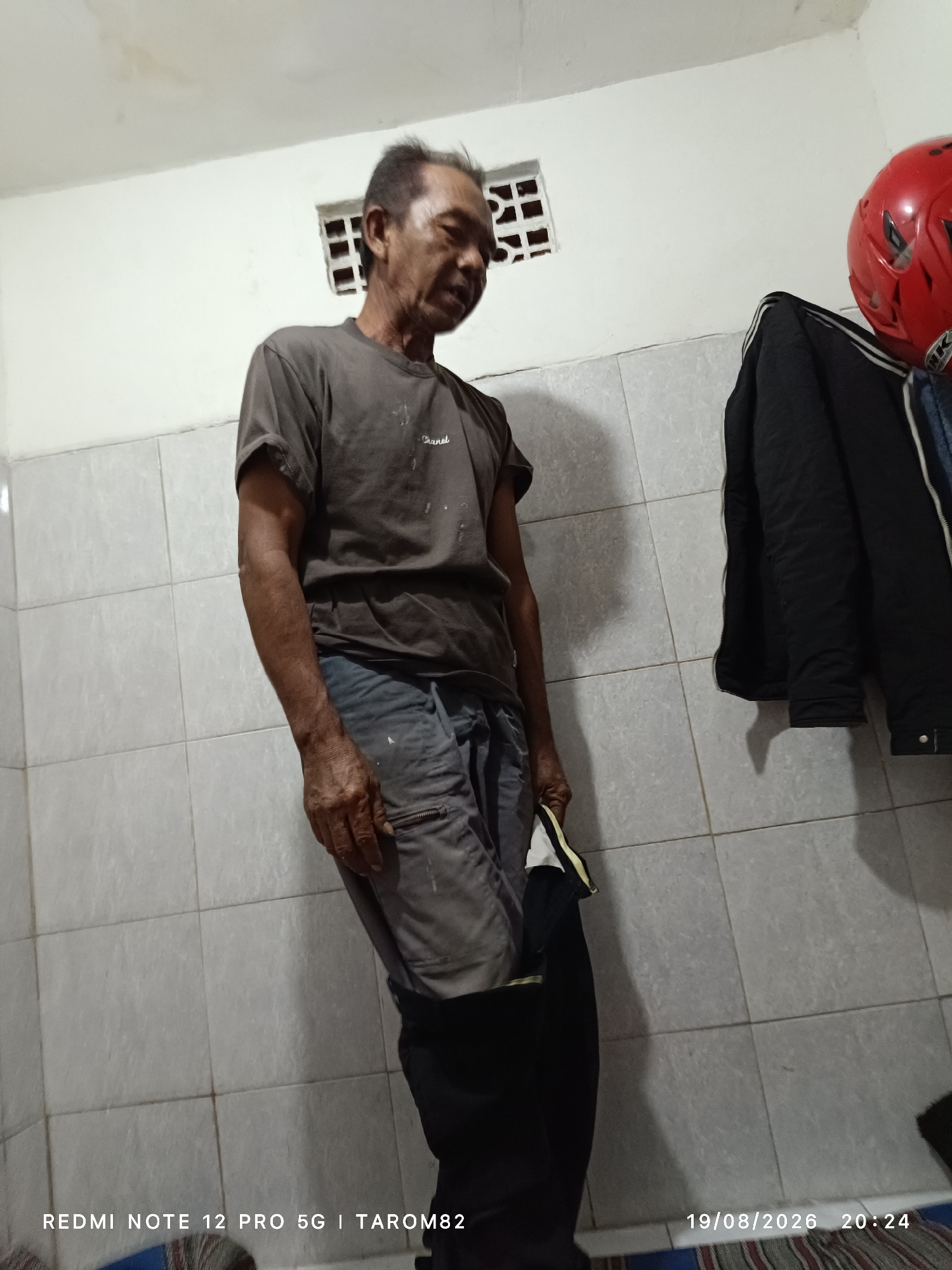 The image depicts an outdoor scene in Sidodadi, Indonesia. An elderly man stands facing to the left, with his head tilted downward and his eyes closed. He is wearing a dark gray t-shirt and gray pants. His right hand is at his side, and he appears to be holding a folded black object with a yellow stripe in his left hand. The man is positioned against a wall covered in light-colored square tiles. To his right, a dark jacket is hanging on a hook. Above the man, a red helmet is visible. The background includes a white wall with a small, perforated vent. The lighting in the scene is dim. The bottom of the image displays text that includes "REDMI NOTE 12 PRO 5G | TAROM82" and the date "19/08/2026 20:24".