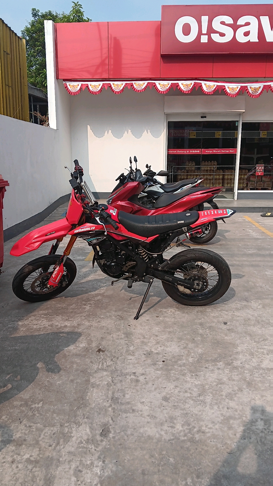 The image shows two motorcycles parked on a concrete surface outside a store with a red awning. The store has a large white sign that reads "o!save" in white lettering. Below the awning, there is a row of red and white flags with a gold emblem. The store entrance has glass doors, and inside, shelves stocked with products are visible. The motorcycle in the foreground is red and black with a dirt bike style. The motorcycle behind it is red and appears to be a scooter. The scene is lit by sunlight, casting shadows on the ground. The store is likely a convenience store or minimarket. The presence of "o!save" suggests it is part of a chain. The red and white flags with the gold emblem are likely Indonesian national symbols, indicating the location is in Indonesia. Given the prevalence of such stores in Indonesia and the specific branding, this scene is consistent with a location in Depok, Indonesia.