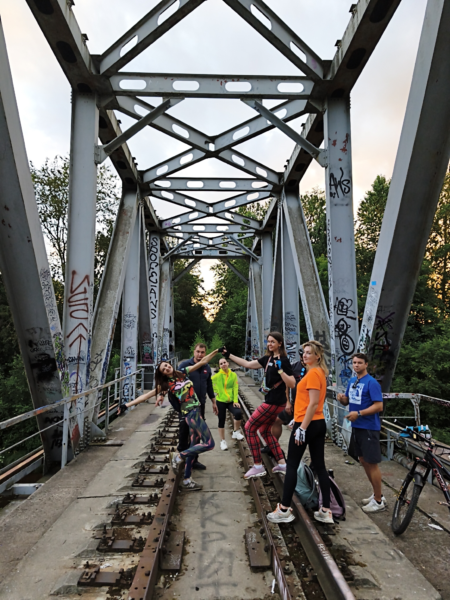 Six individuals, three men and three women, are gathered on an old, graffiti-covered metal railway bridge, possibly in Saint Petersburg, Russia. The group appears to be posing for a photograph, with some individuals gesturing playfully. The bridge is elevated, with trees and a sky with soft clouds visible in the background. The scene suggests an outdoor recreational activity, possibly involving cycling, as a bicycle is parked on the right side of the bridge. The overall atmosphere is casual and energetic.